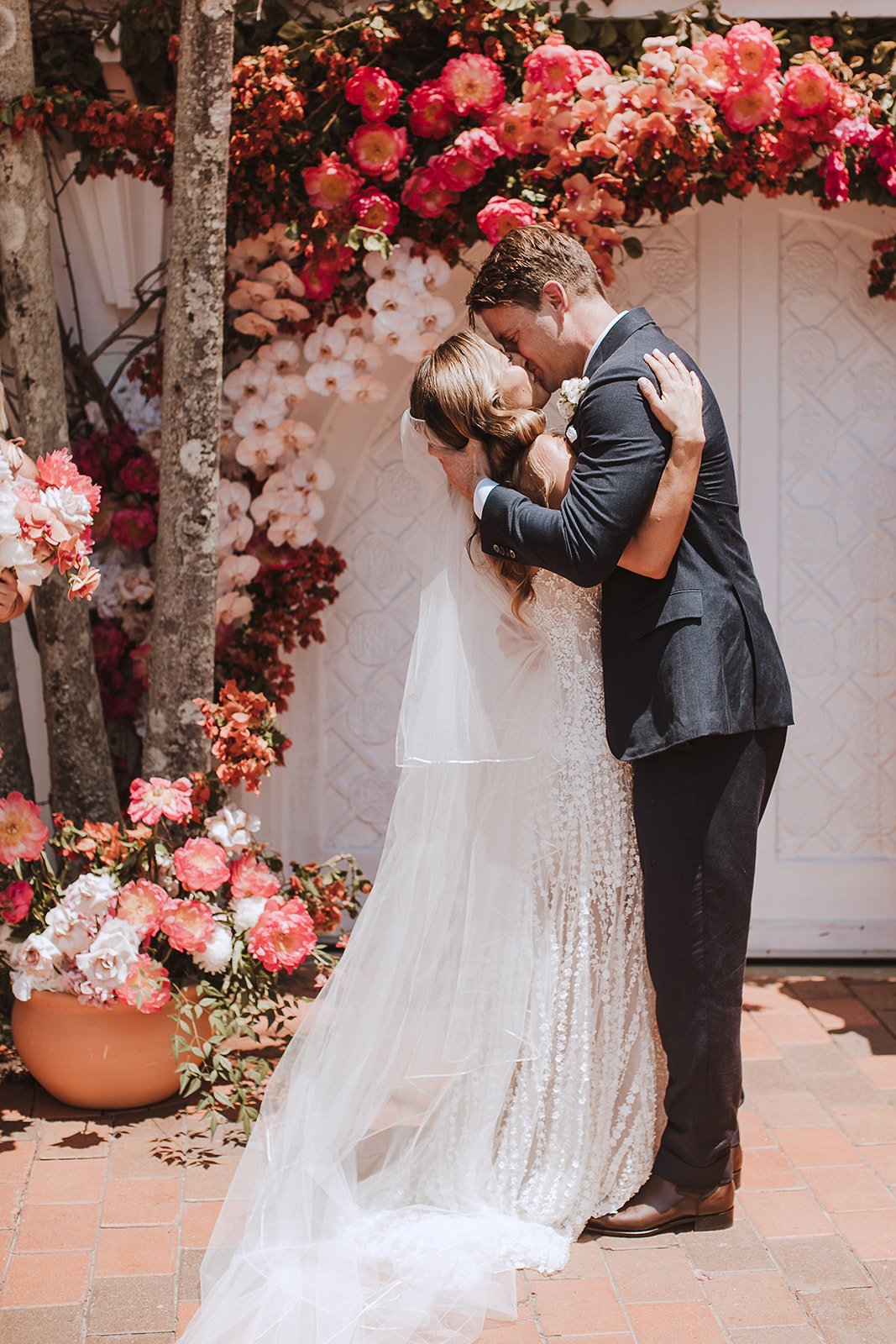 A bride and groom share a kiss during their wedding ceremony, surrounded by pink and white flowers with a decorated backdrop.