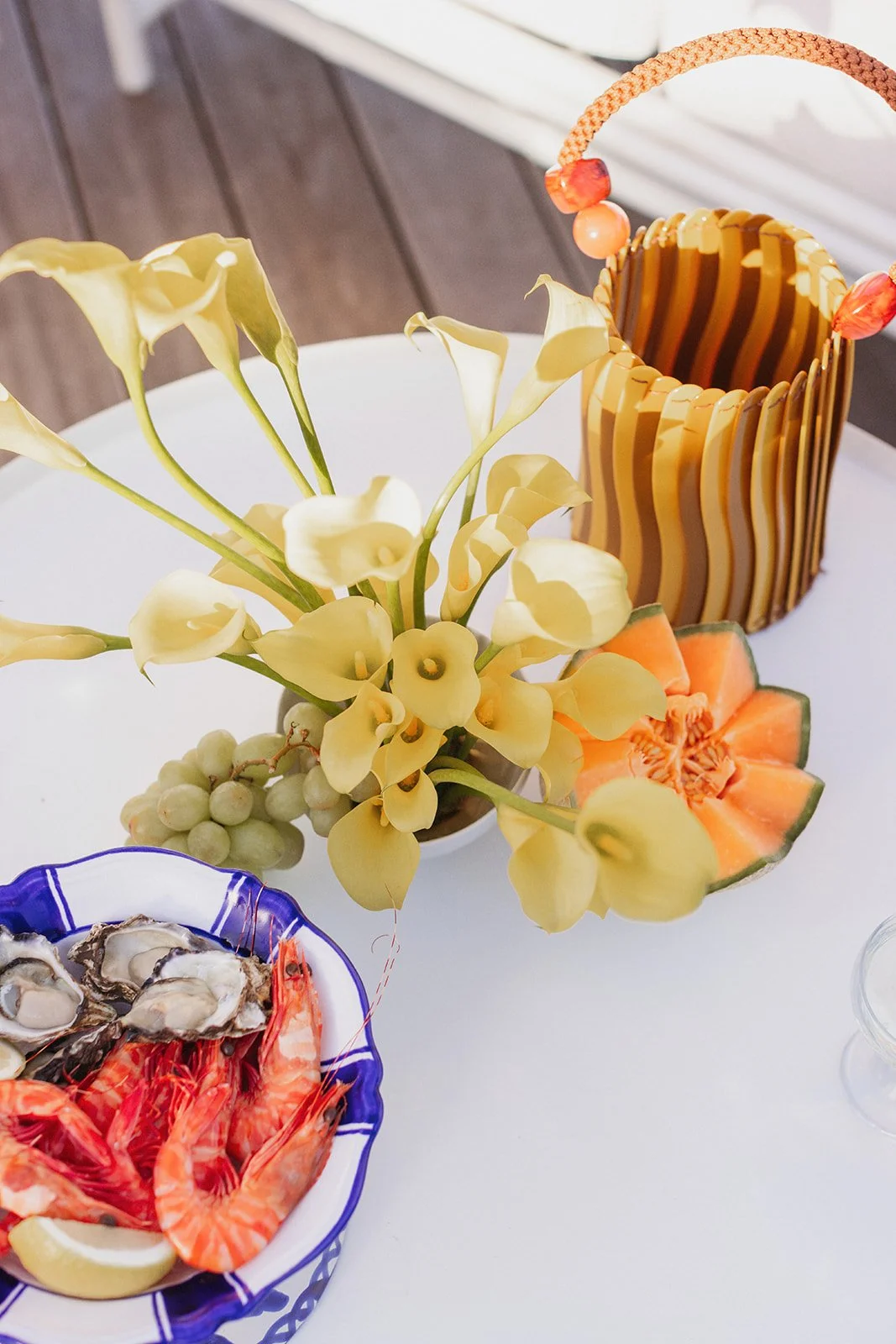 A table with a vase of yellow calla lilies, a plate of oysters and shrimp, a piece of cantaloupe, and a wicker basket.