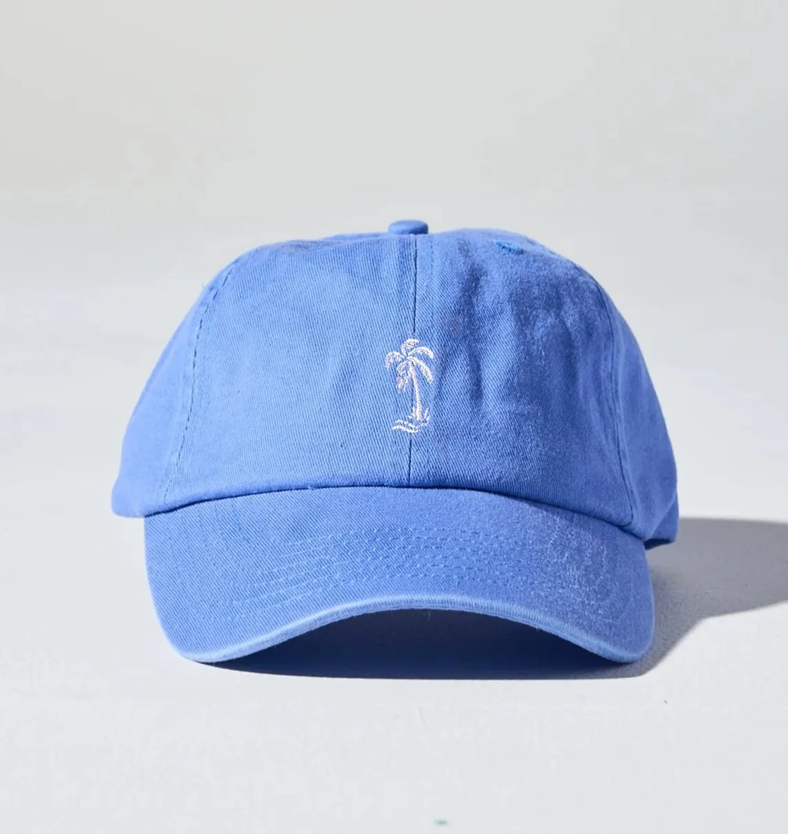 A light blue baseball cap with a small white embroidered palm tree on the front, placed on a white surface against a plain background.