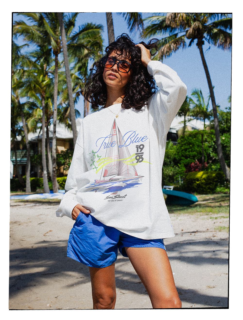 Woman with curly hair wearing sunglasses, a white sweatshirt with a sailboat graphic and text, and blue shorts, on a beach with palm trees and a blue kayak in the background.