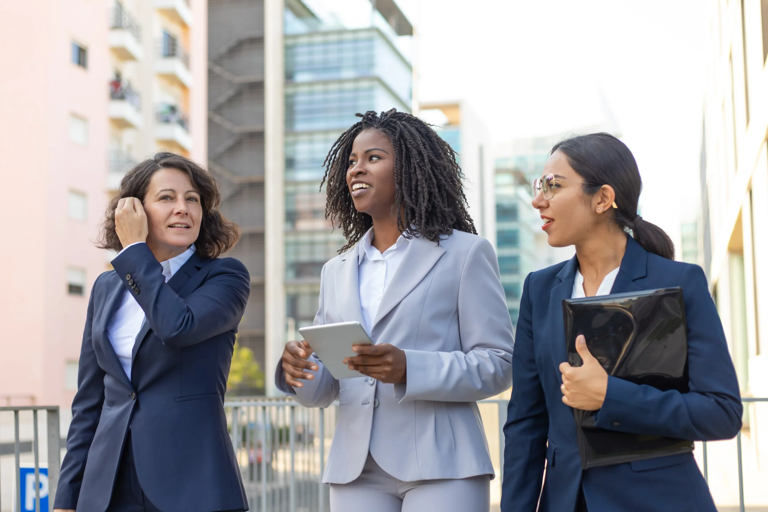 Three businesswomen in suits having a conversation outdoors in an urban setting, with tall buildings in the background.