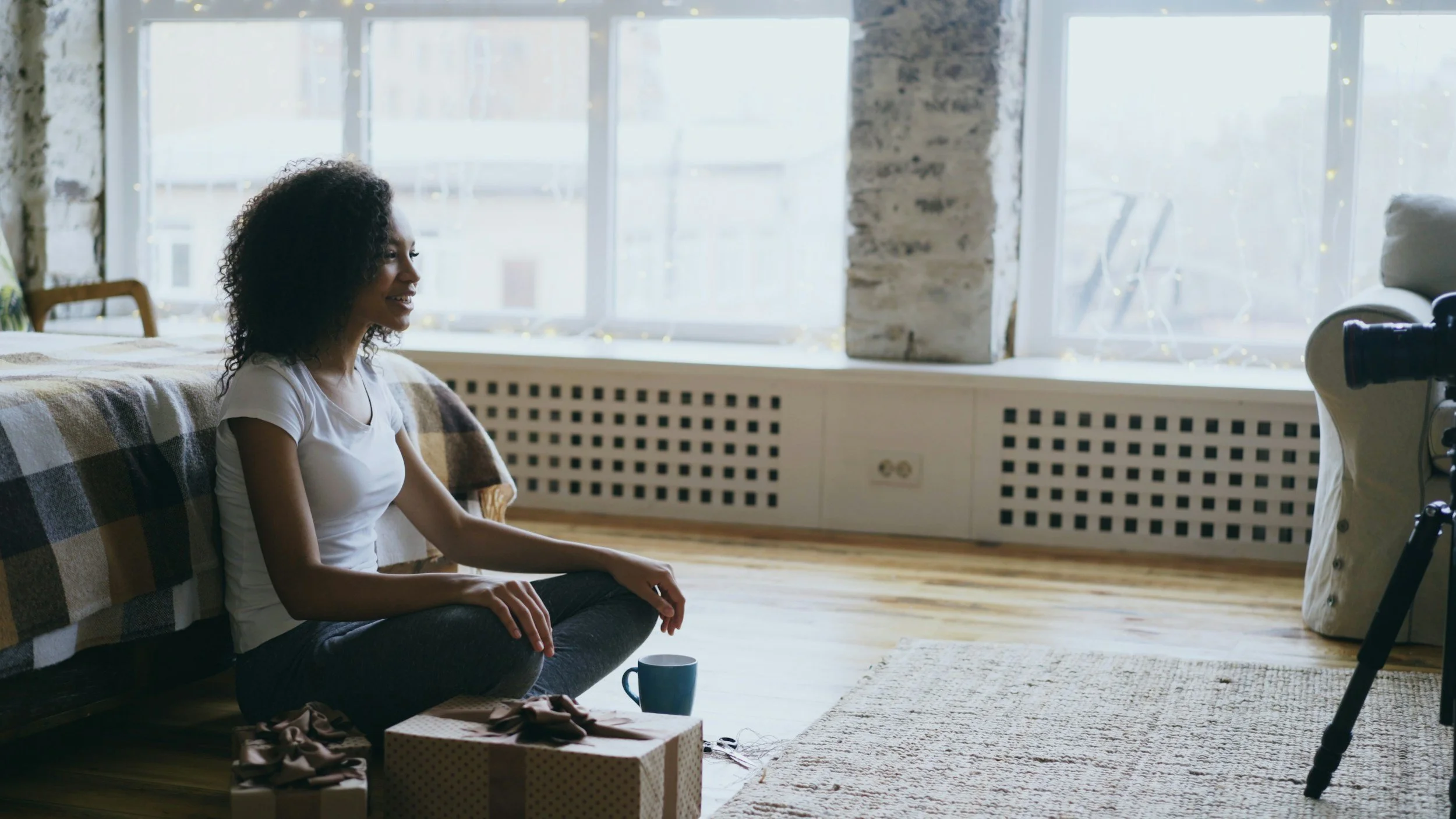 A woman sitting cross-legged on the floor in front of a window, smiling, with a gift box and a mug nearby, likely preparing to record or participate in a video.