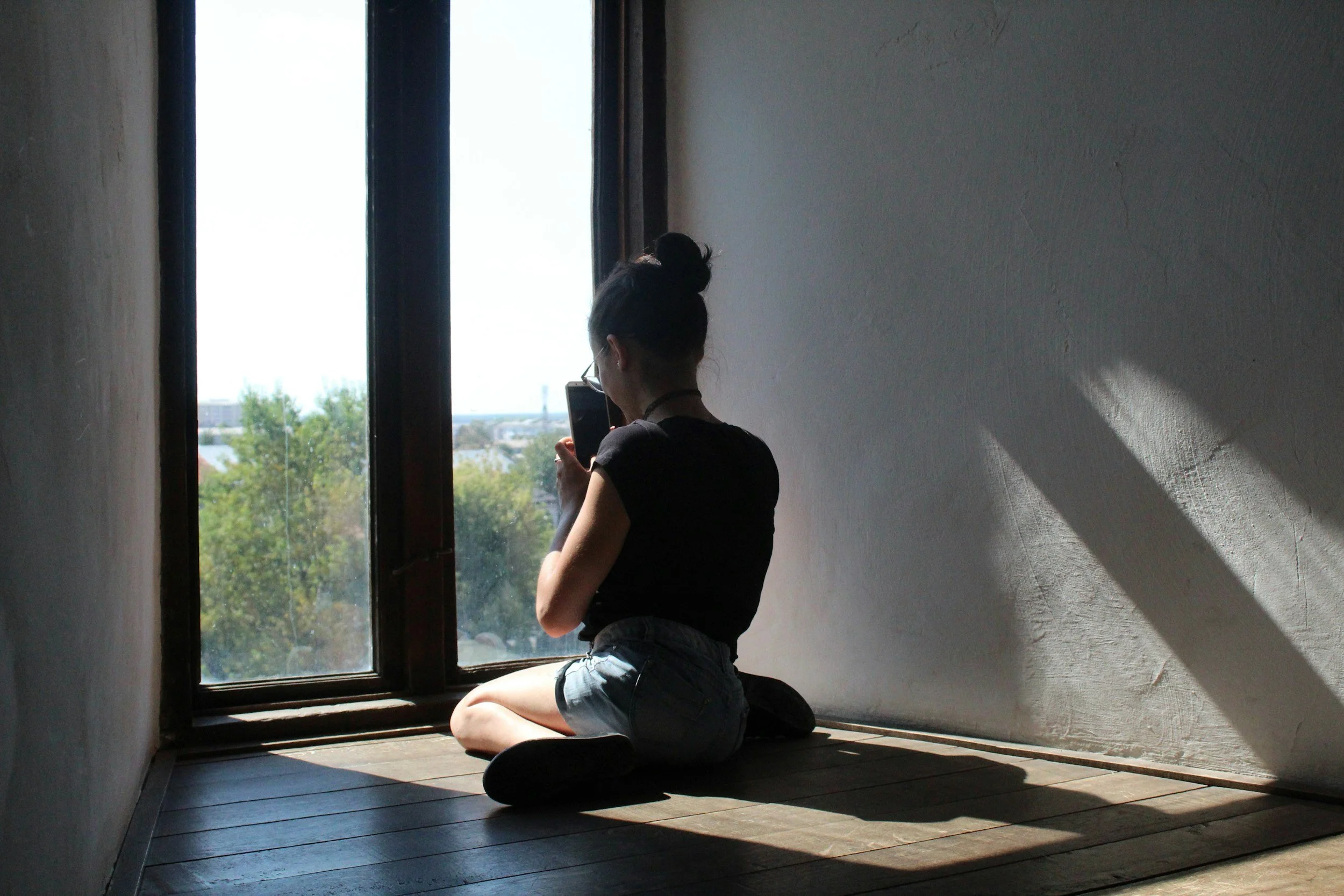 Young woman with a top bun hairstyle sitting on wooden floor in front of large window, taking a photo with her phone. Sunlight casts shadows inside the room.