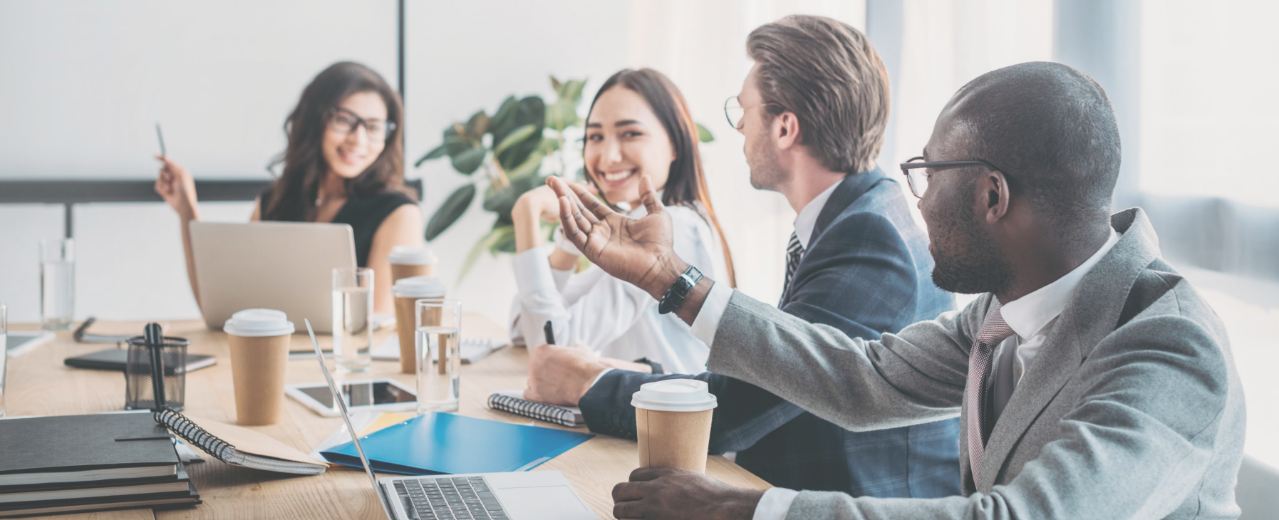 Business meeting with four diverse professionals talking and smiling at a conference table with laptops, notebooks, coffee cups, and a plant in the background.