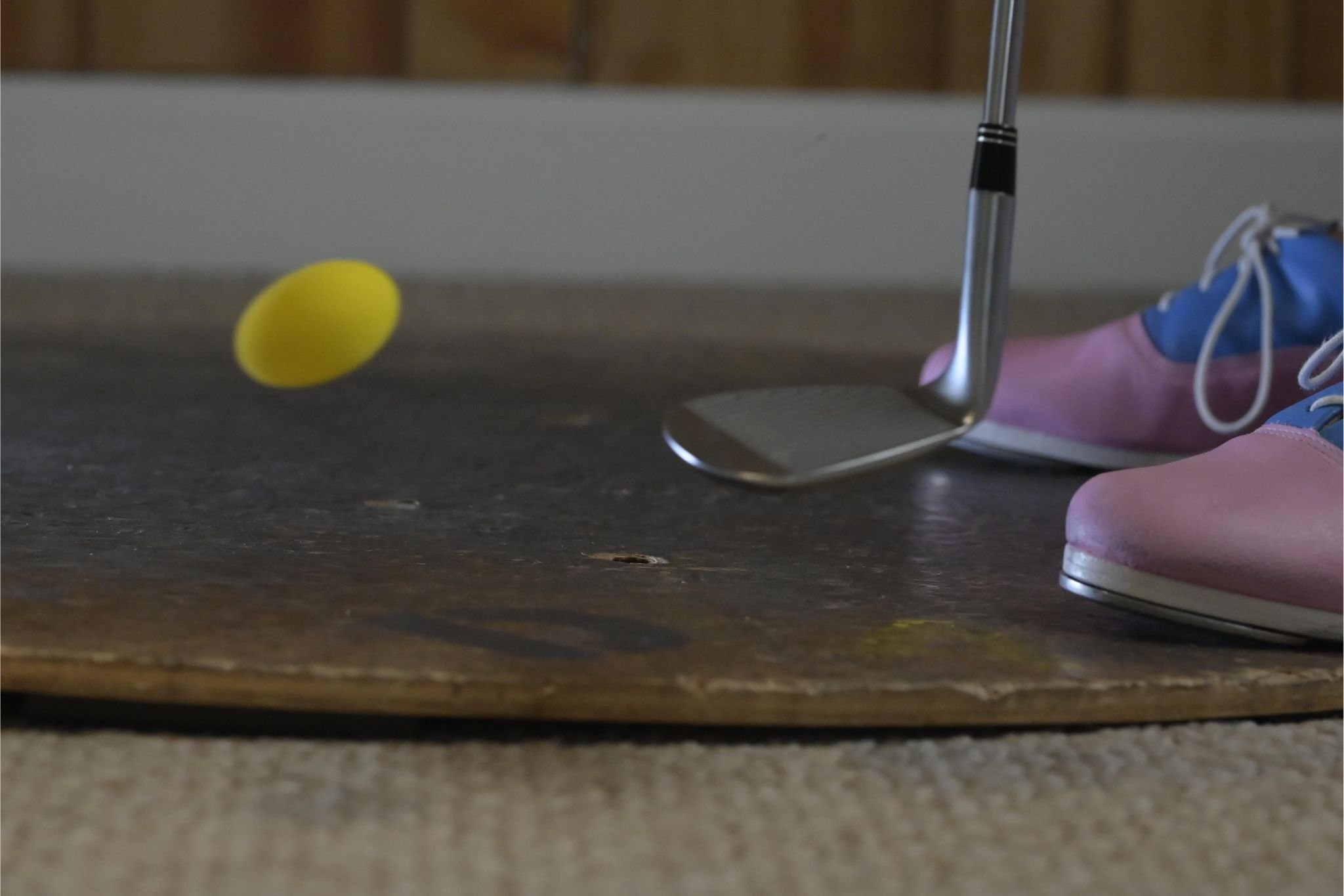 A yellow golf ball near a metal golf club on a wooden teeing area with pink and blue sneakers in the background.