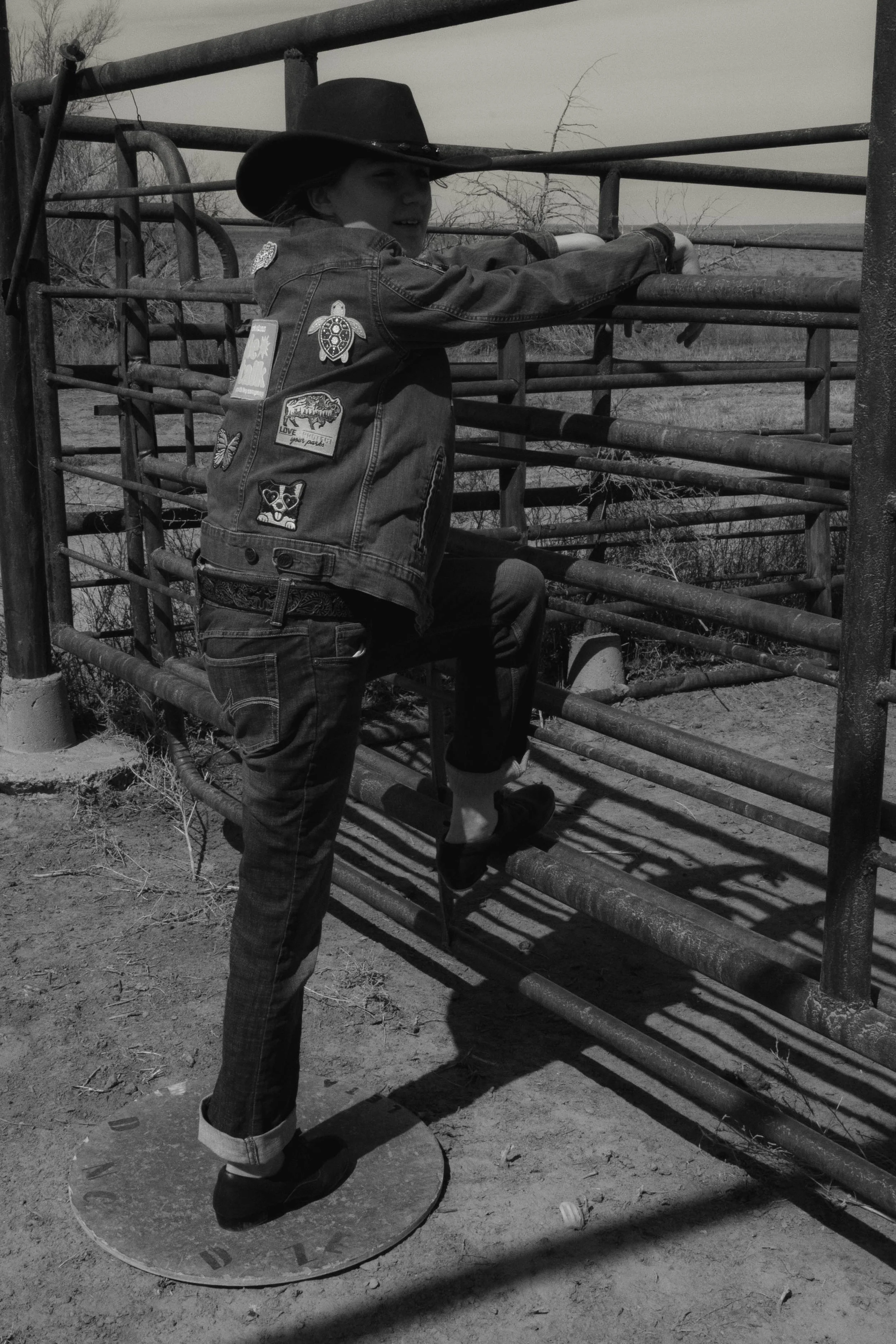 A girl dressed in a cowboy hat and denim jacket with patches, holding onto a metal fence while balancing on a circular platform outside, in a rural setting.