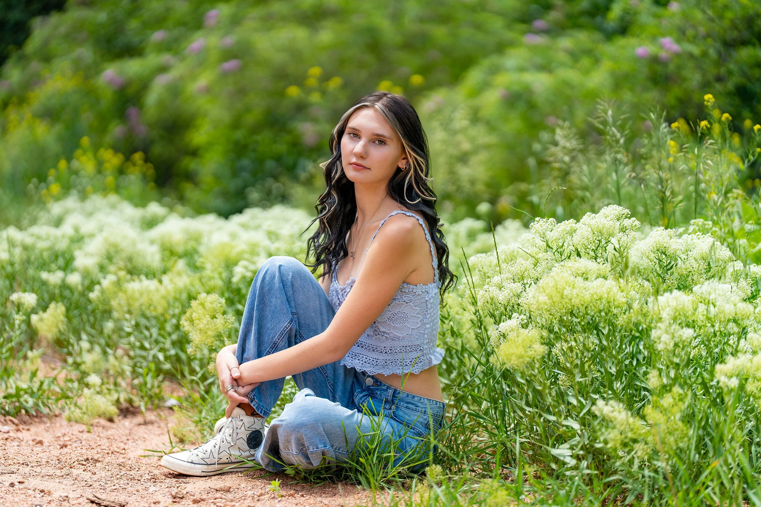 Young woman sitting outdoors on a dirt path, surrounded by white and green flowering plants with greenery and trees in the background.