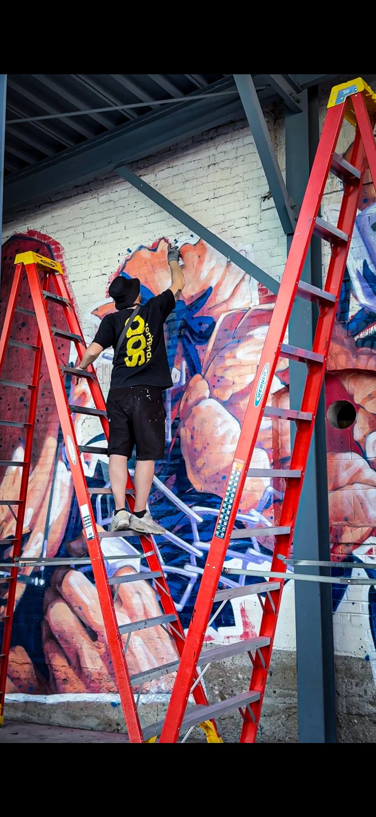 A muralist painting a colorful graffiti mural on a brick wall in an indoor or covered space in Southern California using a spray can. The artist is standing on a ladder and wearing a black T-shirt, shorts, and a hat.