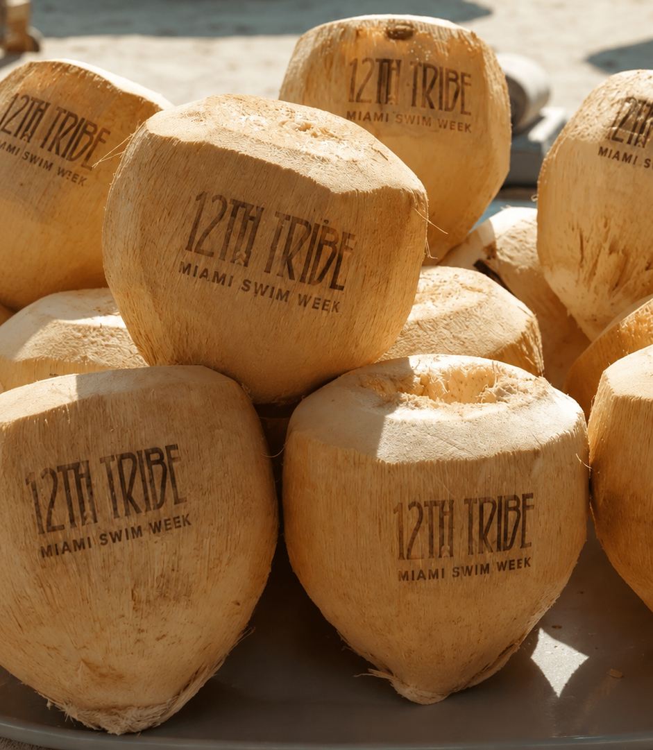 A pile of young coconuts with the logo '12th Tribe Miami Swim Week' printed on them, placed on a surface outdoors.