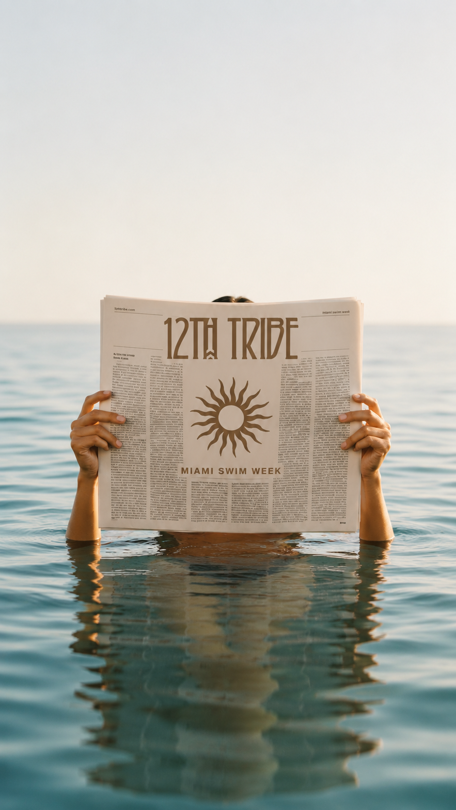 Person holding a newspaper with '12th Tribe' and 'Miami Swim Week' written on it, partially submerged in water, with the ocean and sky in the background.