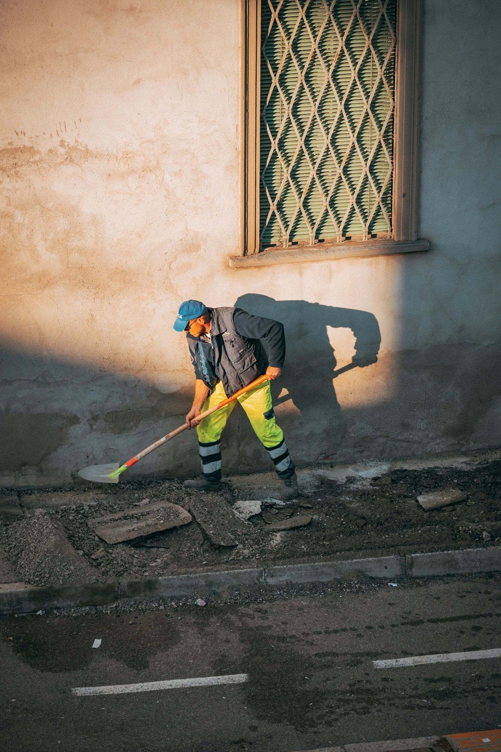 Worker wearing a blue cap and high-visibility clothing, using a shovel to perform road work next to a wall with a barred window.