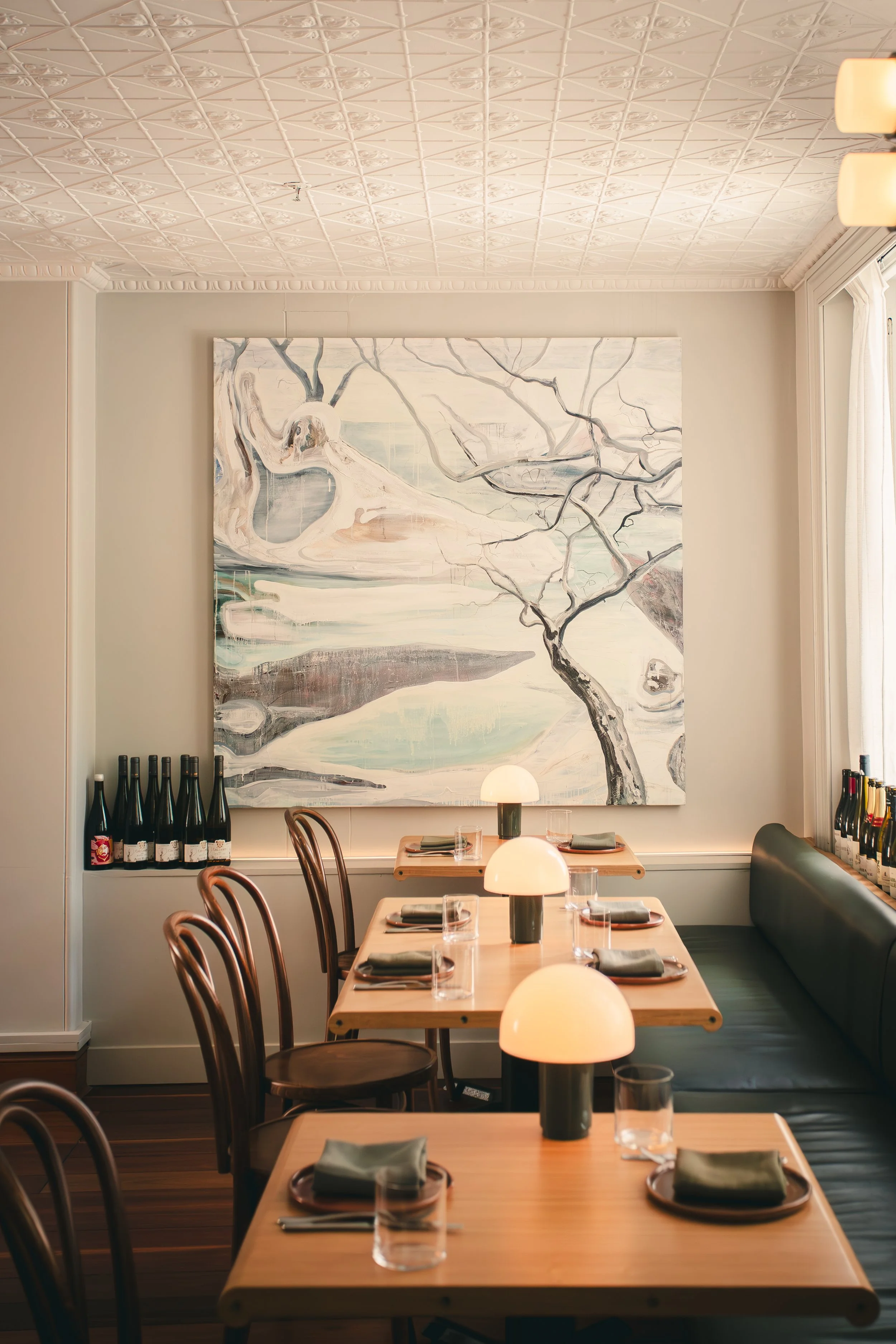Dining area with four wooden tables, each set with napkins, glasses, and lamps, positioned in front of a large abstract painting of a landscape with trees and water. Bottles are on the window ledge and on a shelf near the corner.