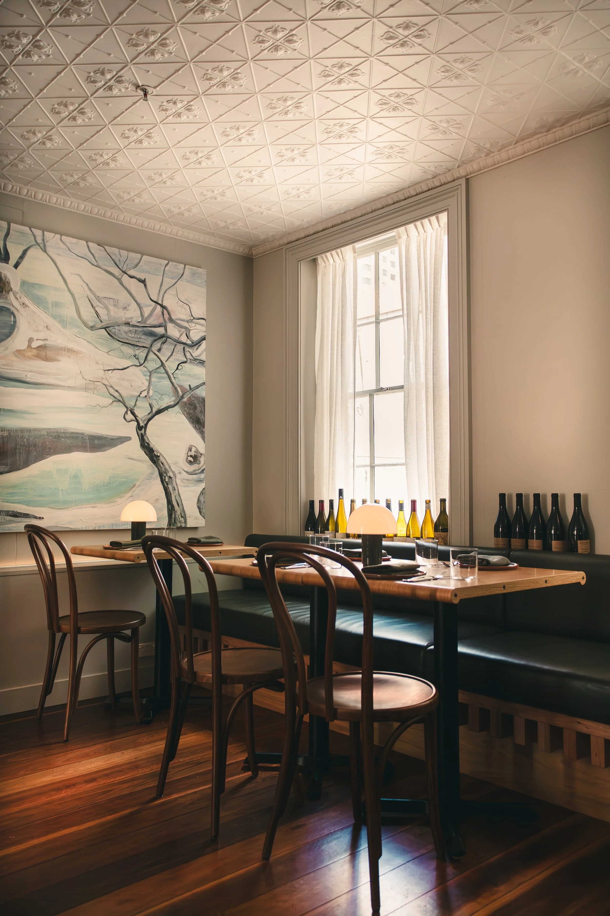 A cozy restaurant dining area with a wooden table set for dining, featuring black cushioned seats, candles, wine bottles along the window sill, and abstract artwork on the wall, illuminated by natural light through a large window with curtains.