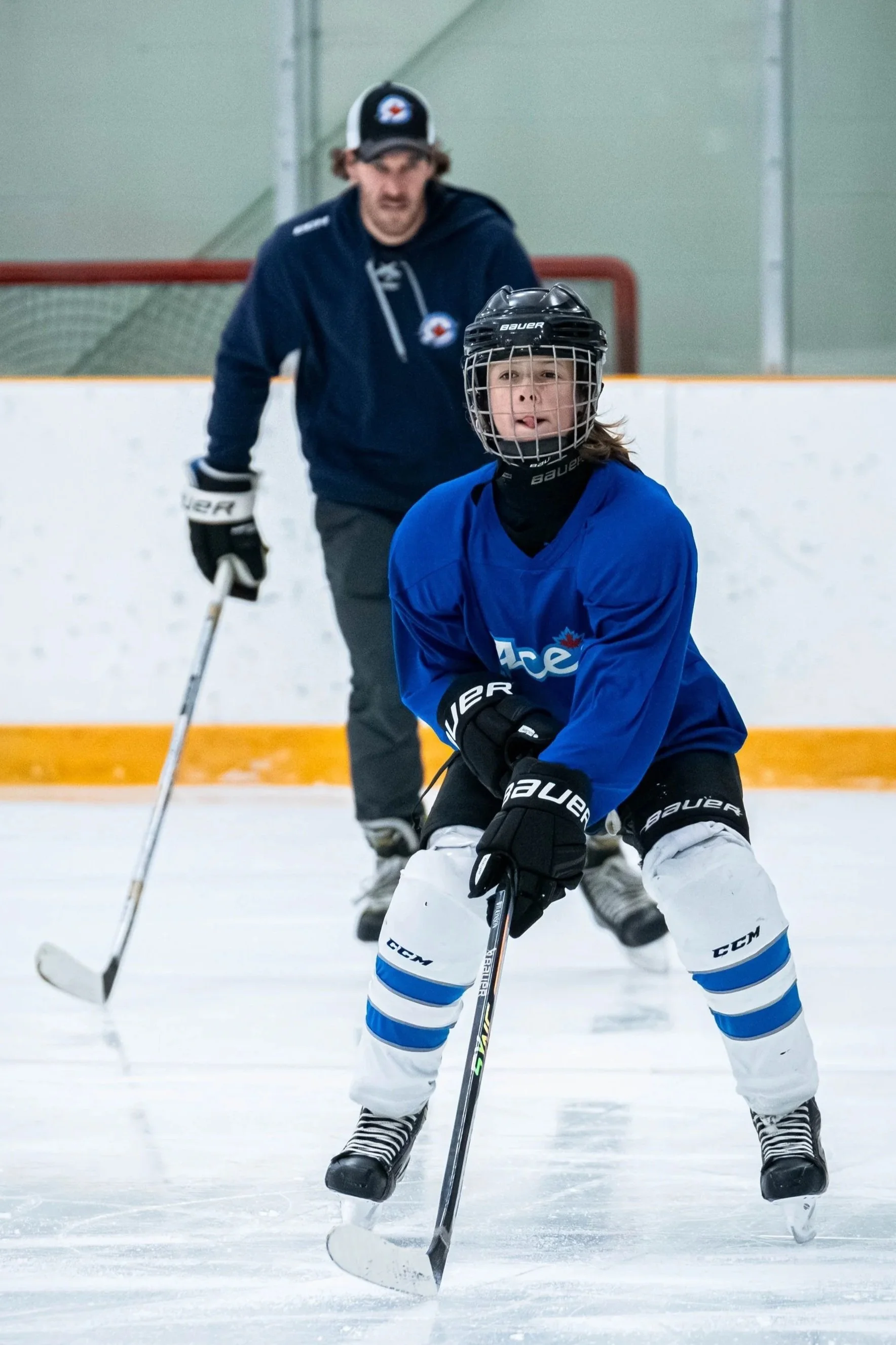 A young hockey player wearing a helmet, blue jersey, and white hockey pants practicing on ice with a coach in the background. working on alignment, balance and control on skates