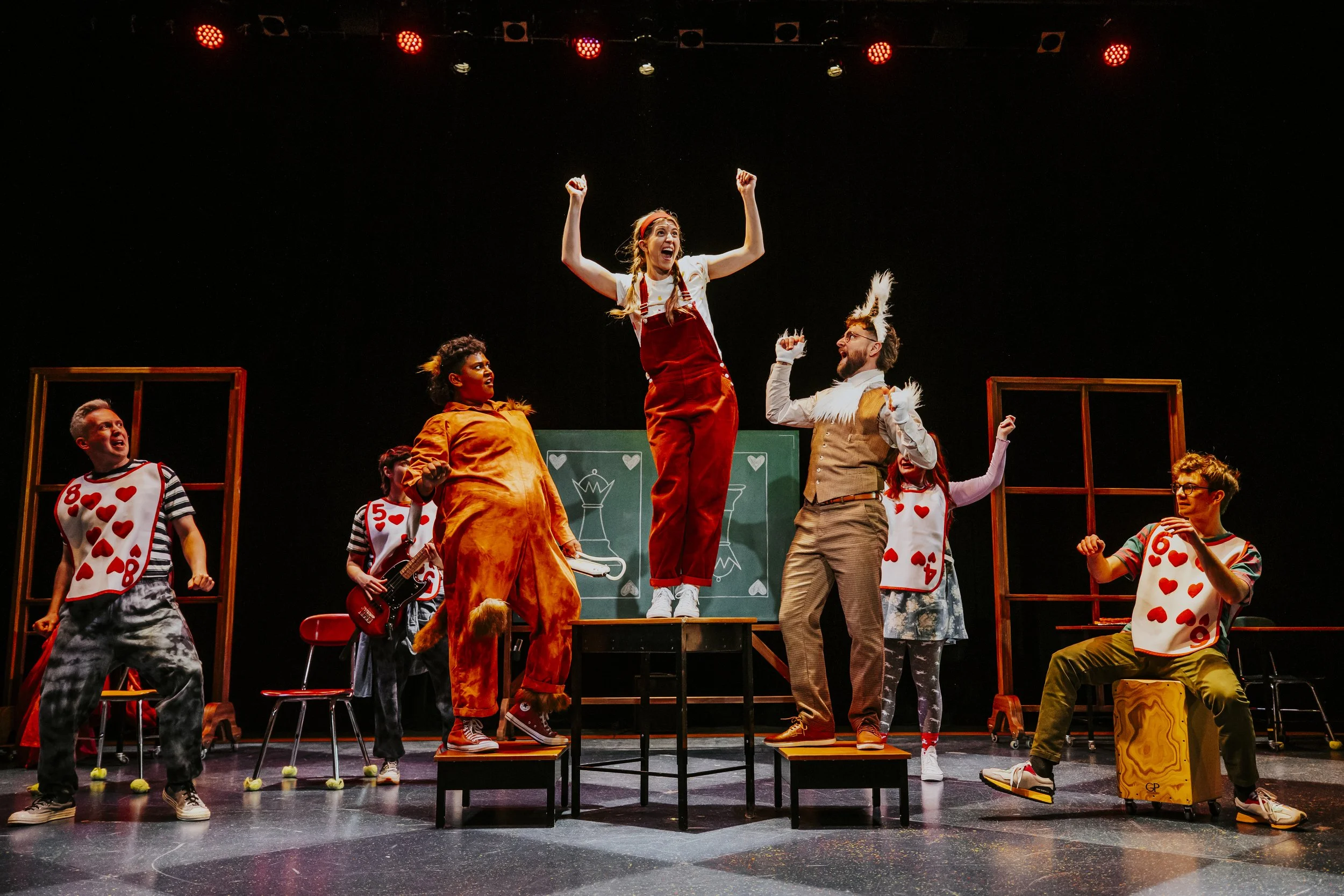 Theatrical performance with six actors dressed in colourful costumes, some wearing playing card-themed outfits, on a stage with red lighting. One actress stands on a platform with raised arms, and others are engaged in animated poses.