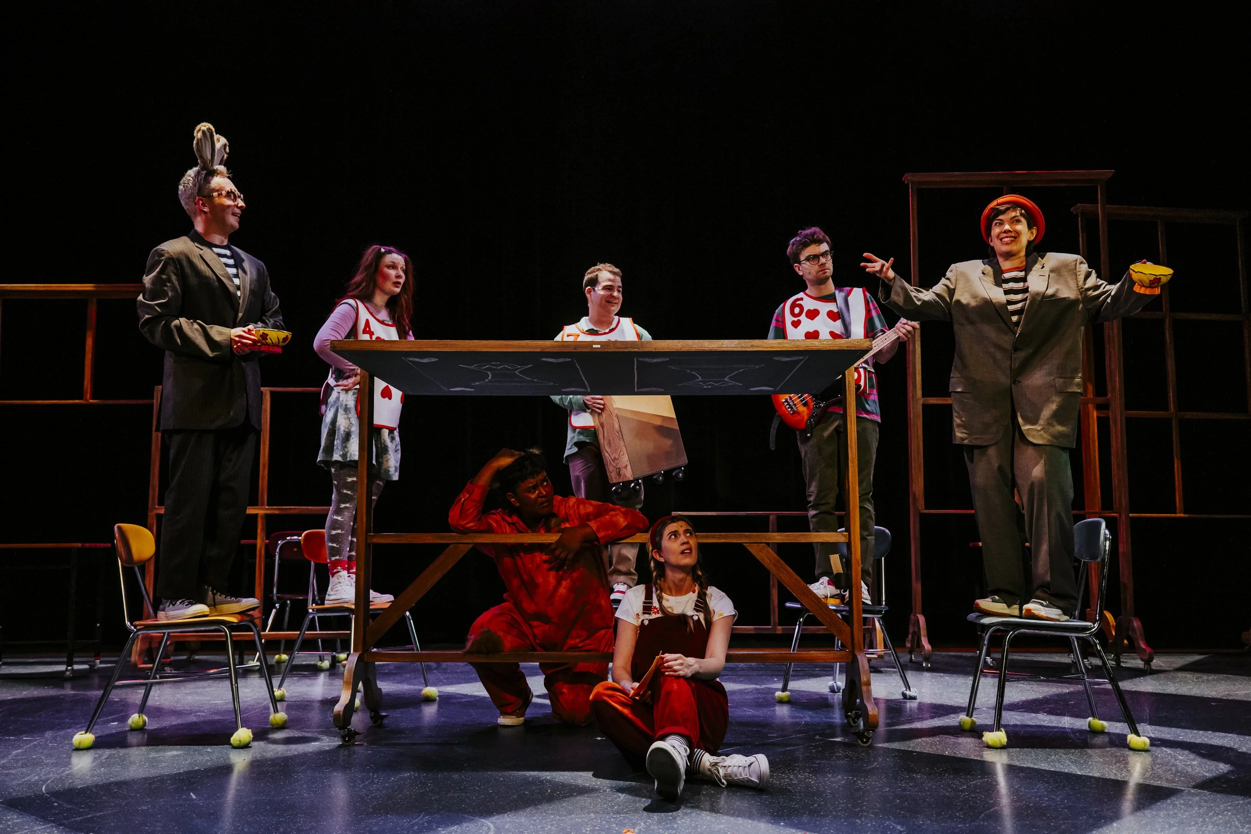 Stage scene of a theatrical play with seven performers, some standing on chairs and tables, others sitting or crouching, dressed in colorful costumes, with expressive gestures under stage lighting against a black background.