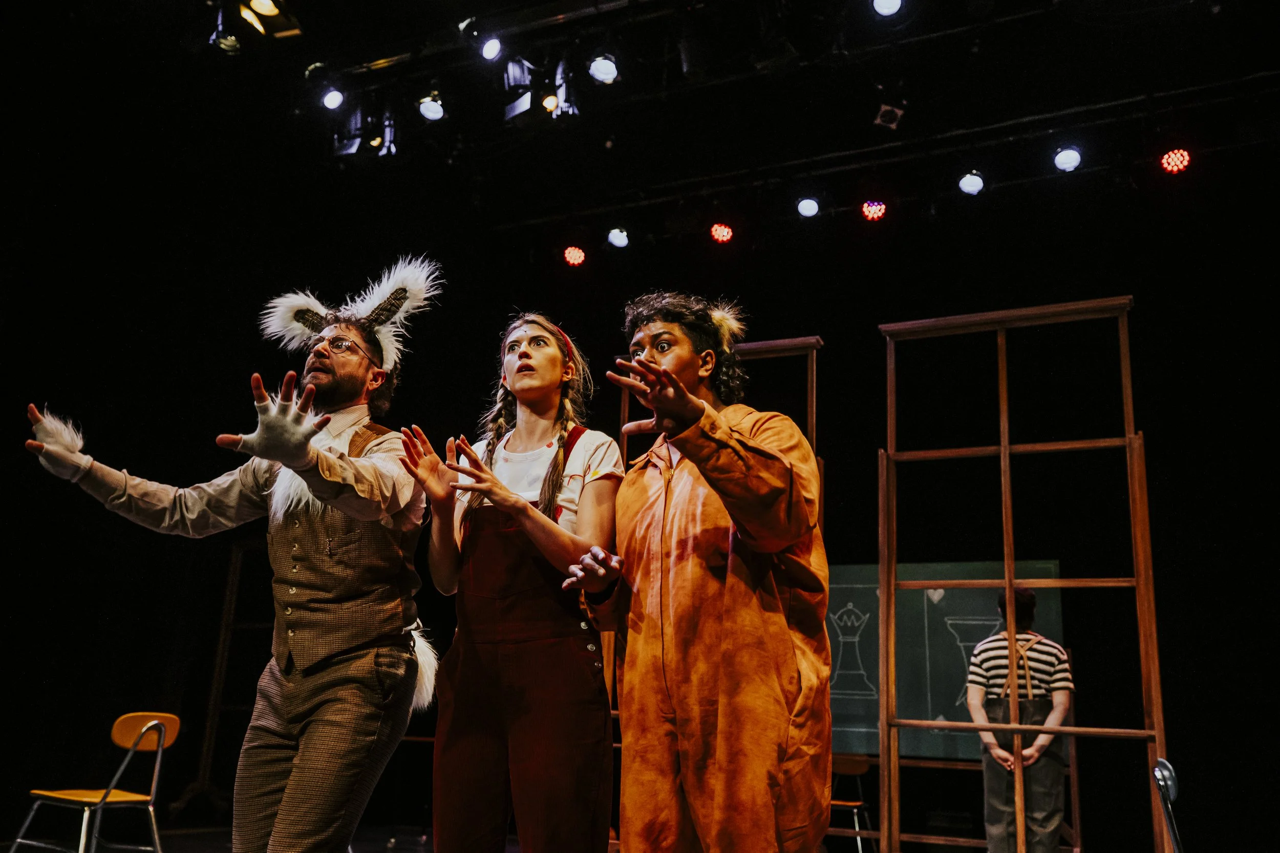 Three actors on stage during a theatrical performance, with one dressed as a cat, another in overalls, and a third dressed as a rabbit, performing in front of stage props including a wooden frame and a chalkboard.