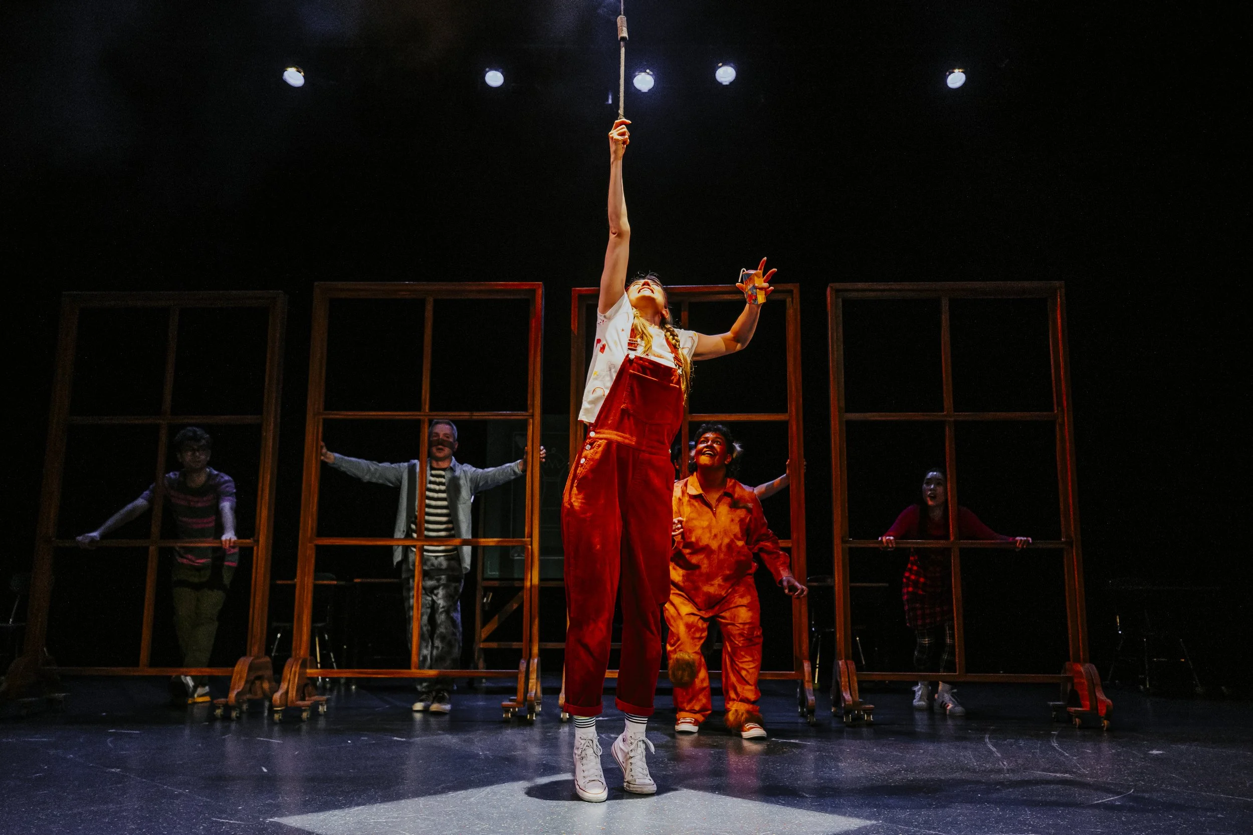 People on stage performing a theatrical play with a woman in overalls and white sneakers reaching upward, and others behind wooden frames, under stage lights.
