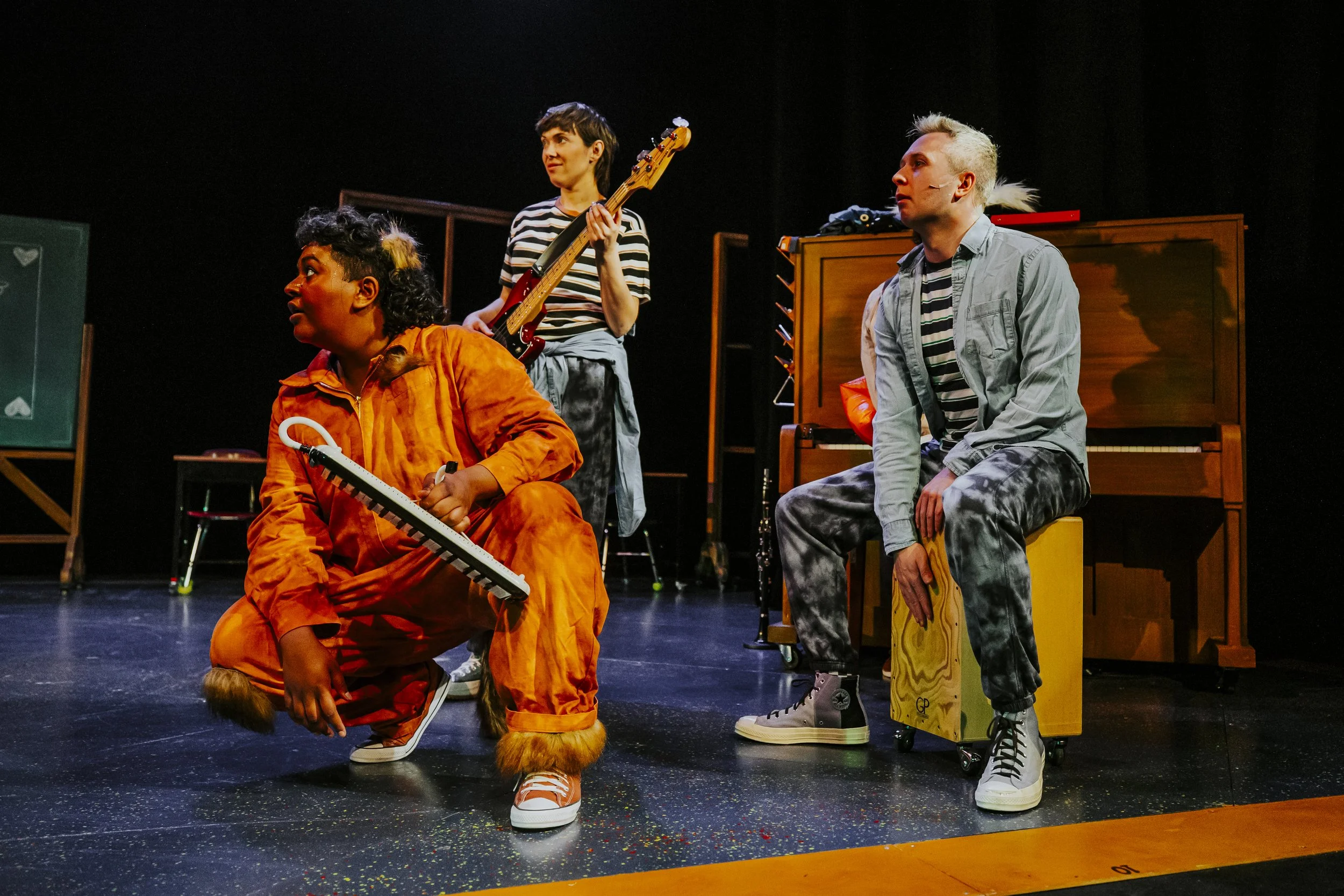 Three performers on stage, one crouching with a melodica, one standing with a bass guitar, and one sitting on a portable cajón drum, all dressed in casual and colourful clothing, with musical instruments and stage equipment behind them.
