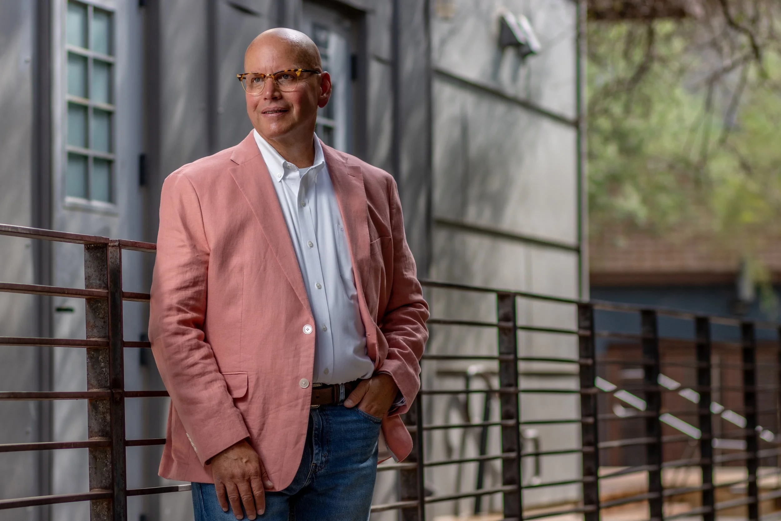 A man wearing glasses and a pink blazer stands outdoors next to a metal railing, with a neutral expression on his face, in front of a gray building with windows and some trees in the background.