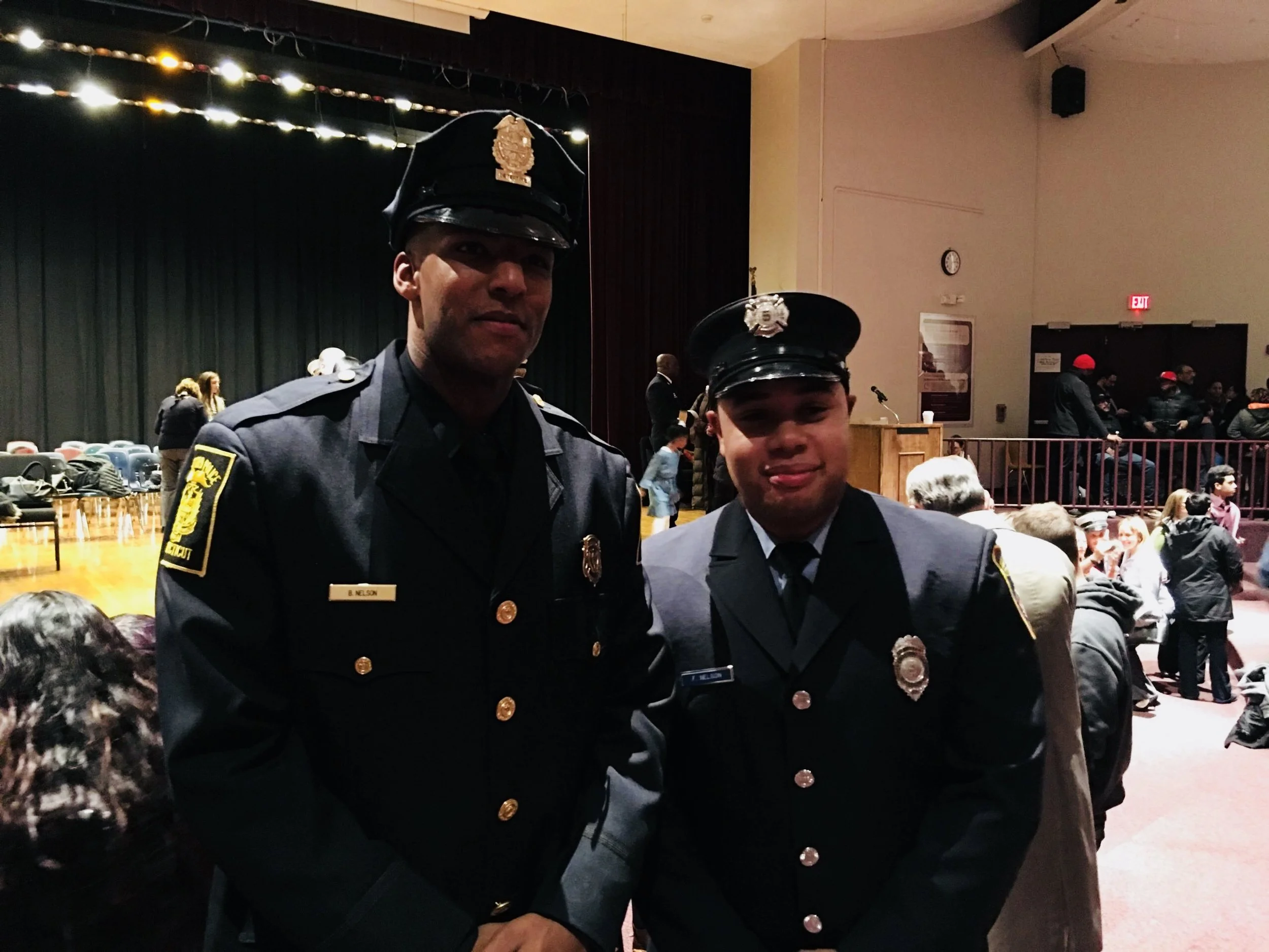 Two uniformed police officers at an indoor event with a stage and audience in the background.