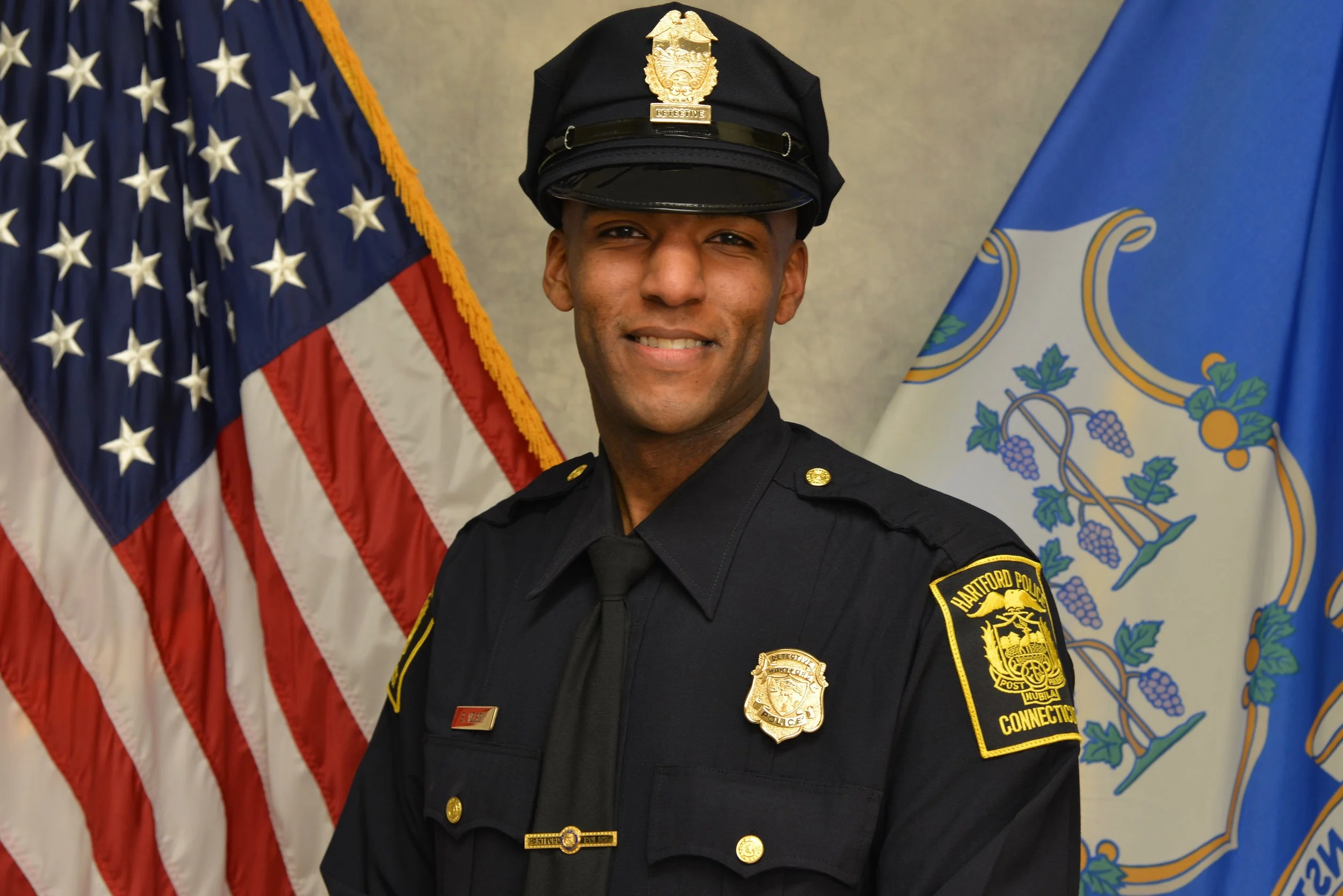 Police officer in uniform standing in front of American and Connecticut flags.