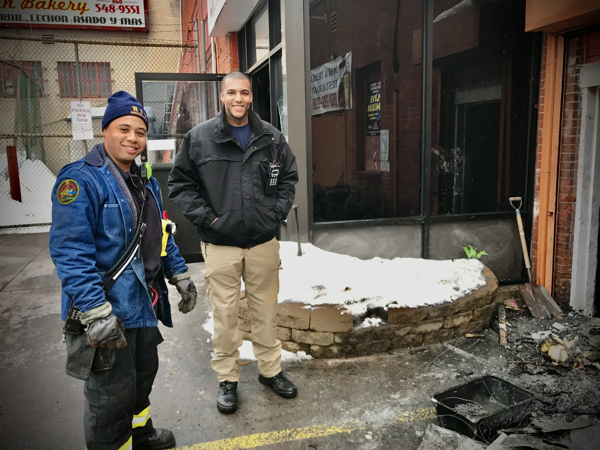 Two men, one in a firefighter uniform and the other in casual attire, stand smiling outside a building. Snow is on the ground, and debris is scattered. There's a shovel leaning against the wall and business signs in the background.