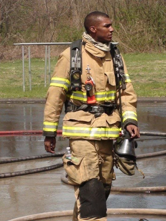 A Windsor, Connecticut Firefighter in full gear walking outdoors, holding a helmet, with fire hoses on the wet ground at the Connecticut Fire Academy in Windsor Locks, Connecticut.