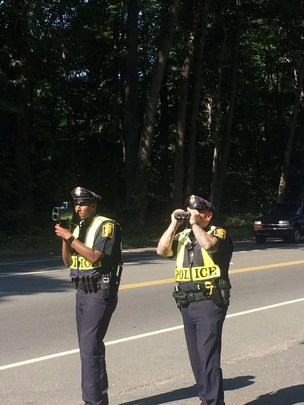 Two Hartford, Connecticut Police Officers, Bryan Nelson and Kevin Nesta, wearing reflective vests are standing by the roadside. One is using a speed gun, and the other is looking through binoculars.