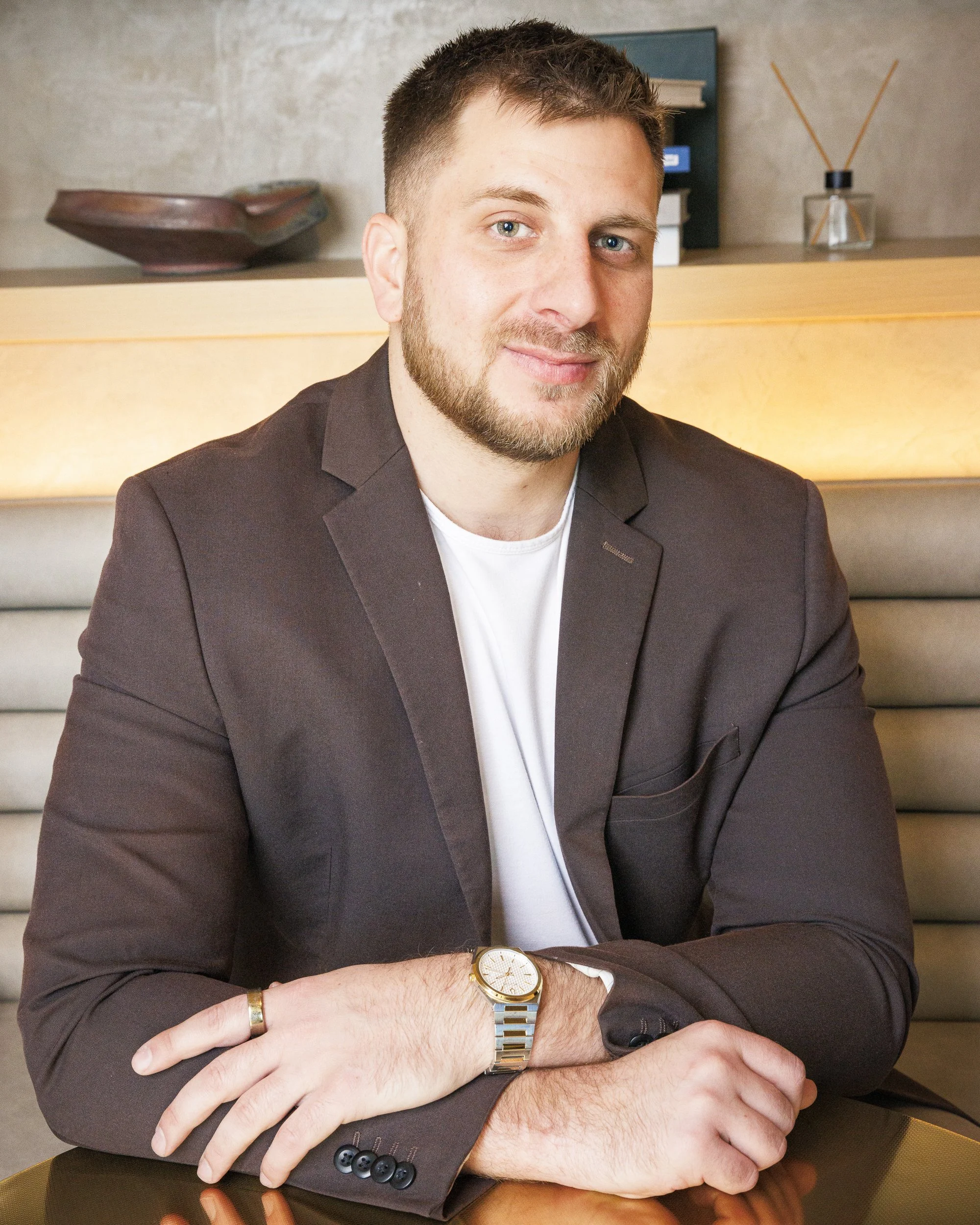 A man with short brown hair and a beard, wearing a dark blazer and a white shirt, sitting at a table with his arms crossed, smiling at the camera in a modern cafe or restaurant setting.