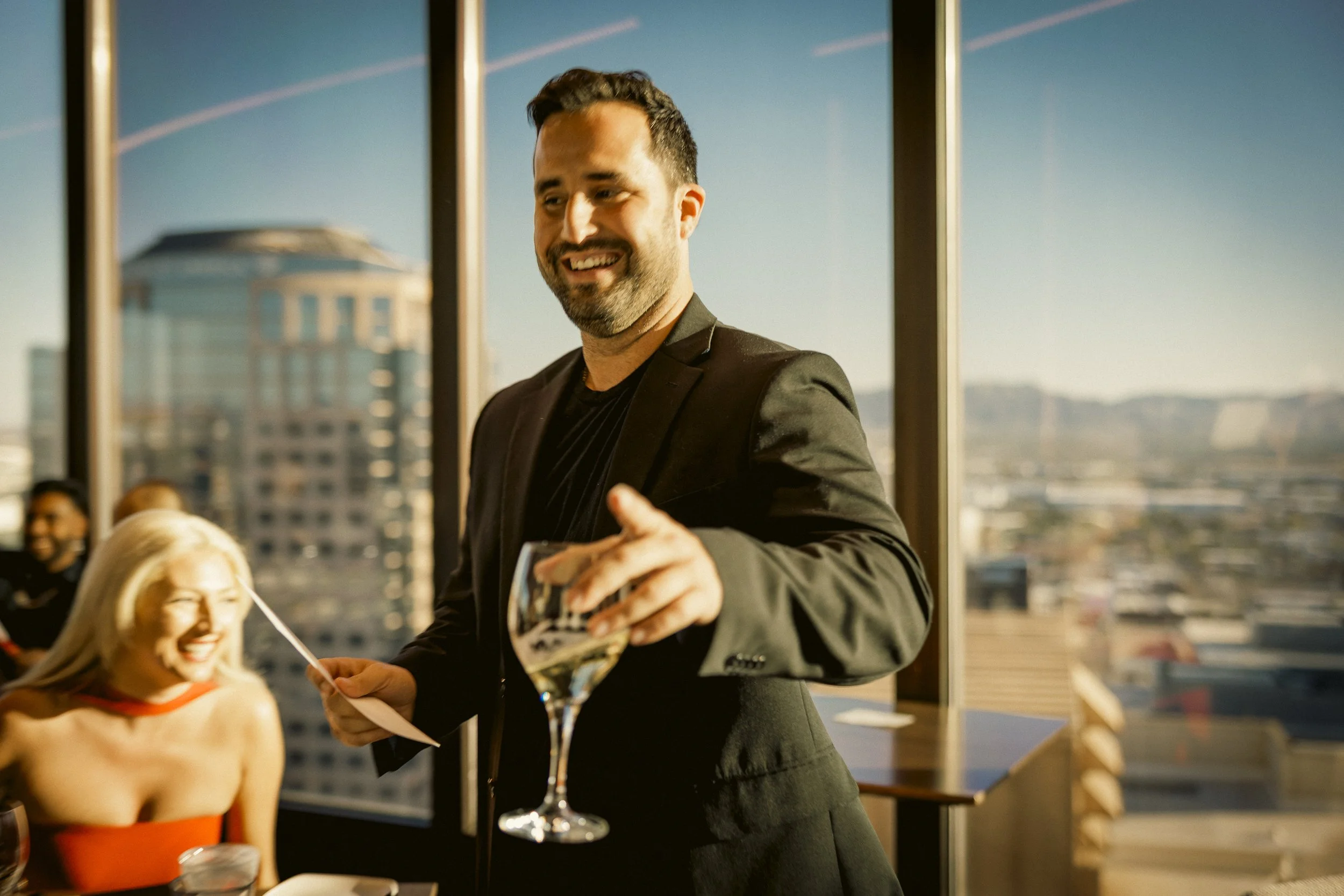 A man with a beard in a black suit smiling and holding a wine glass at a social event, with a woman in a red dress and blonde hair sitting nearby, in a room with large windows and cityscape background.