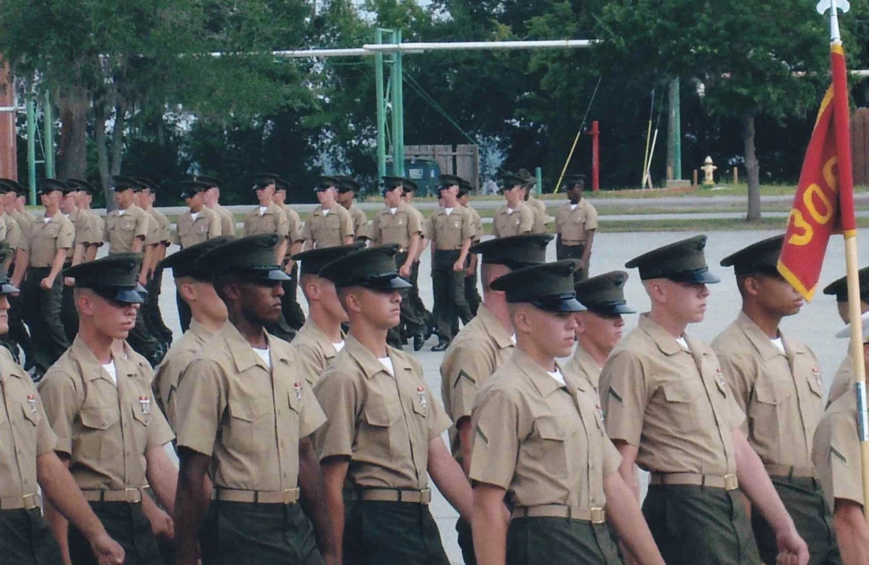 A group of newly graduated United States Marines in uniform marching in formation outside on Parris Island, South Carolina, with the platoon flag held by one person.