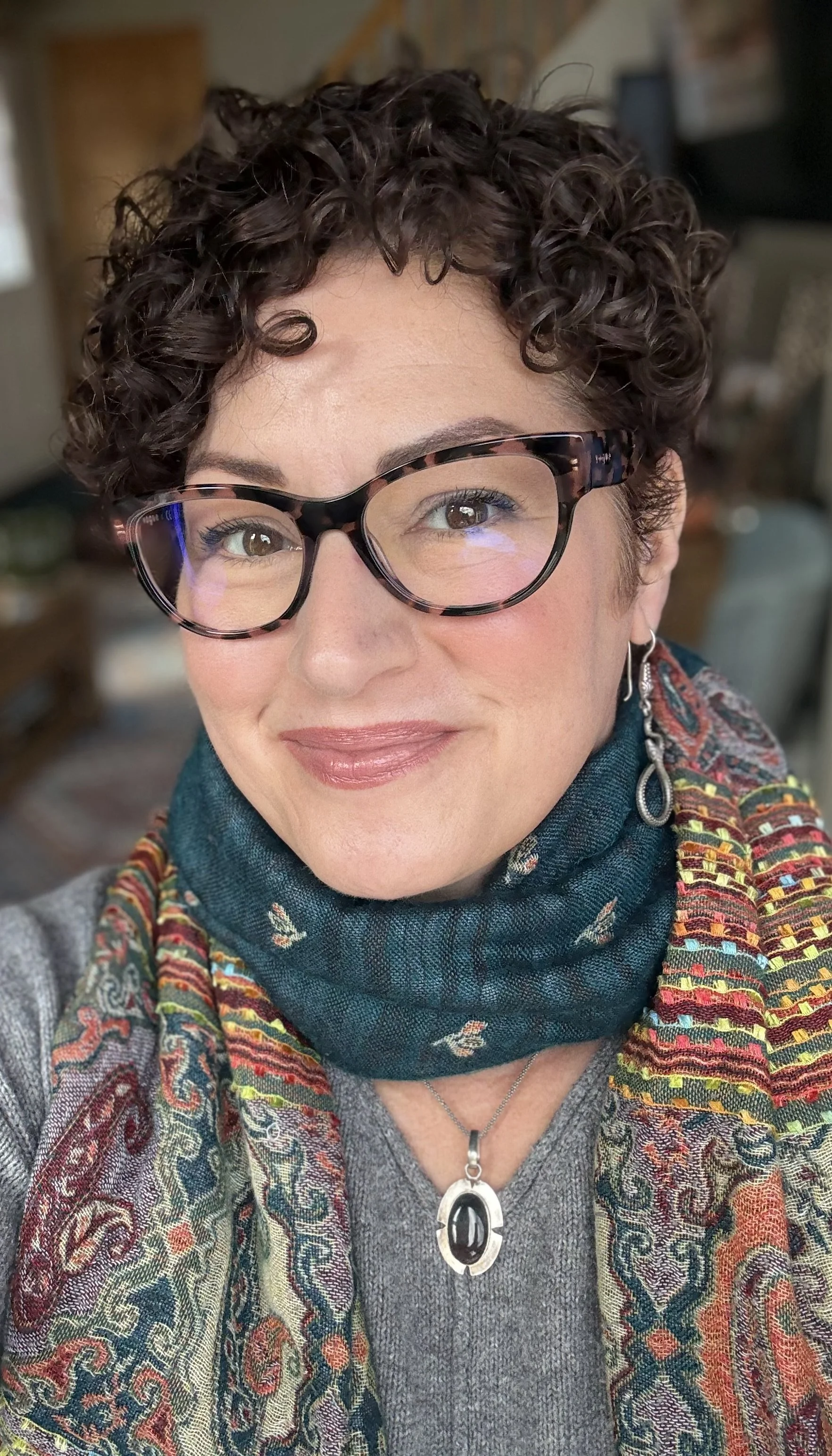 Close-up of a smiling woman with short curly brown hair, wearing glasses, a teal scarf, and a colorful patterned shawl, indoors.