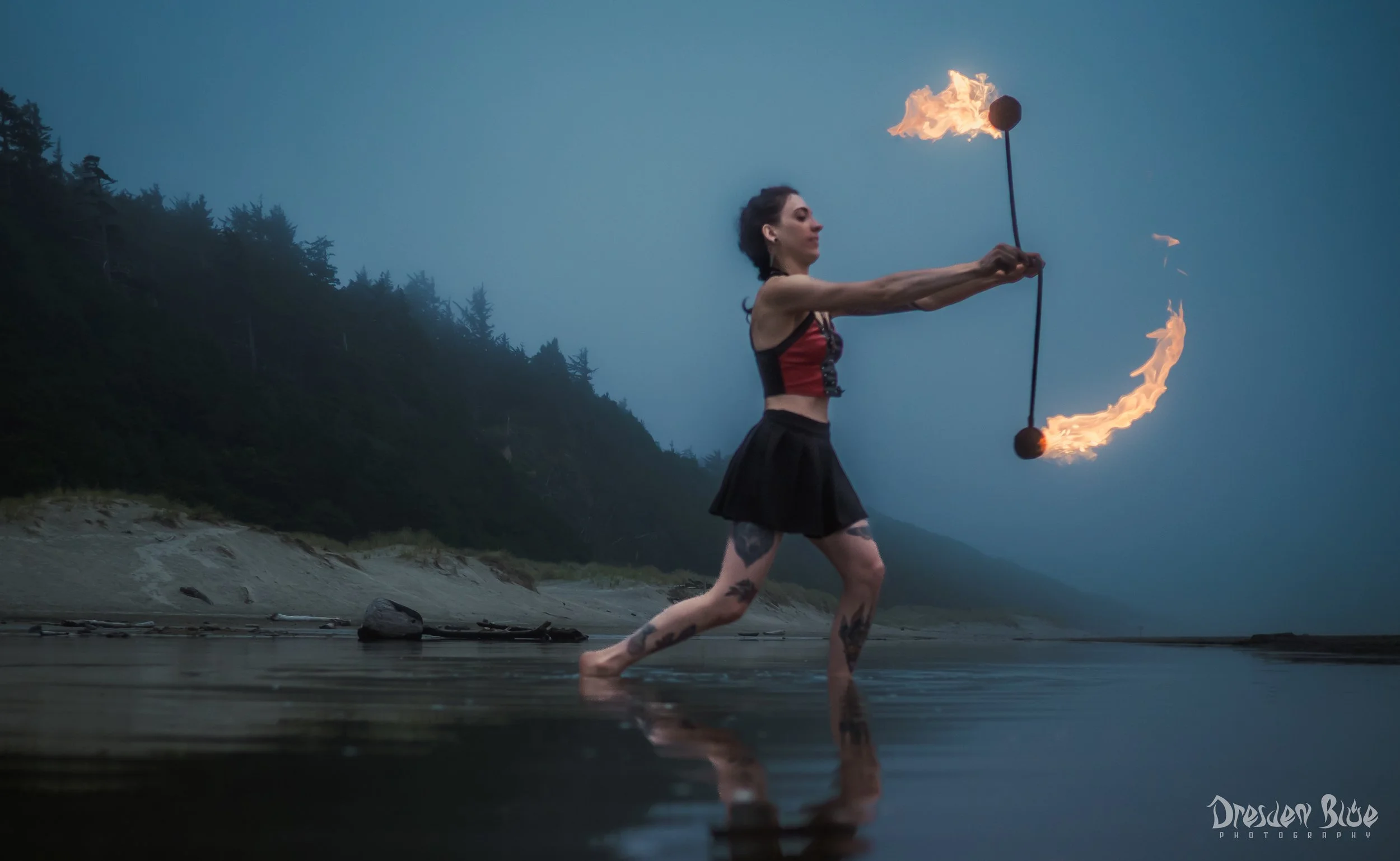 A woman with tattoos on her legs spinning flaming fire poi on a beach at dusk, with mountains and trees in the background.