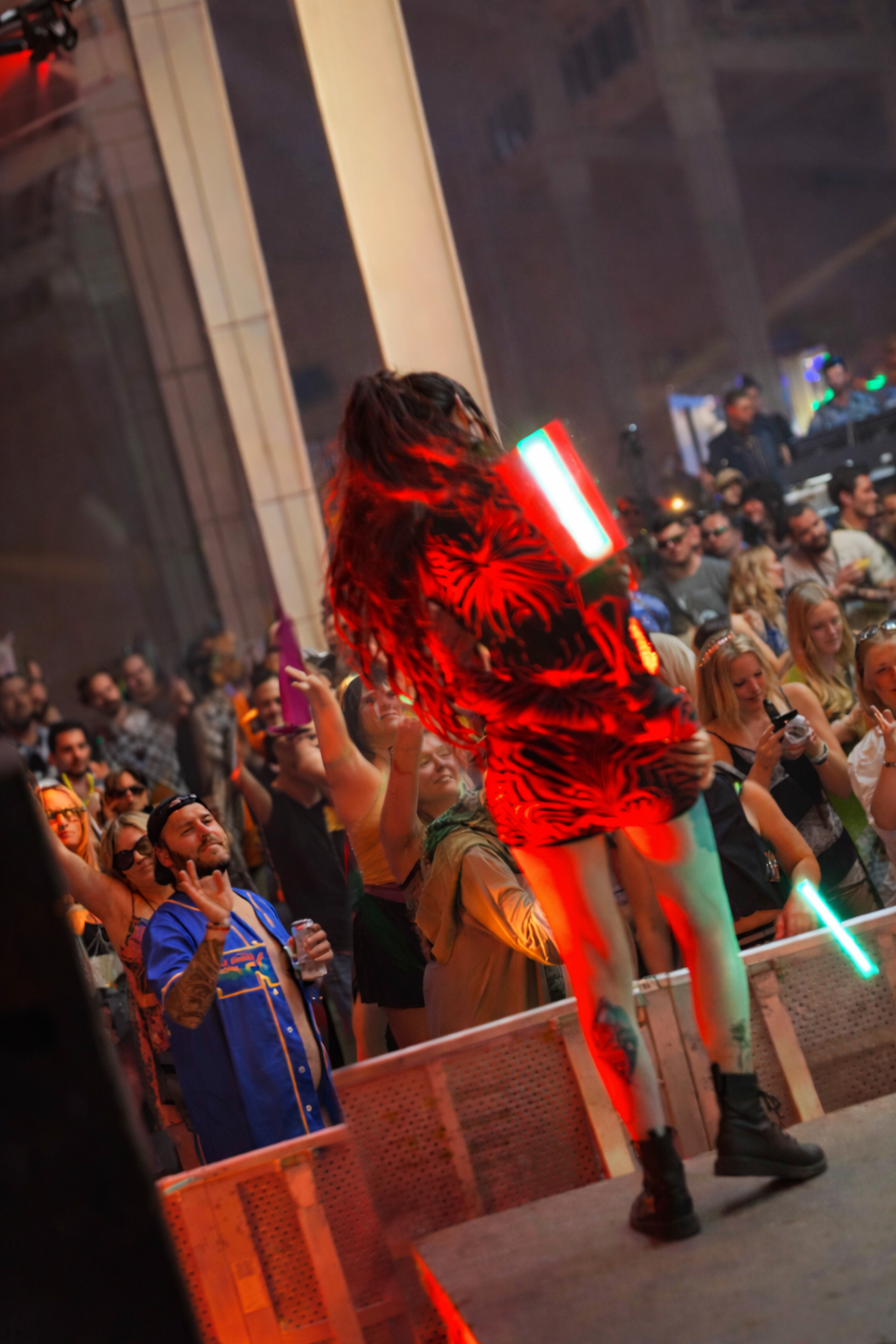 A performer with long hair wearing a vibrant red and black zebra-print outfit and boots, holding a light-up object, performing on stage in front of a large crowd at a lively concert or festival.