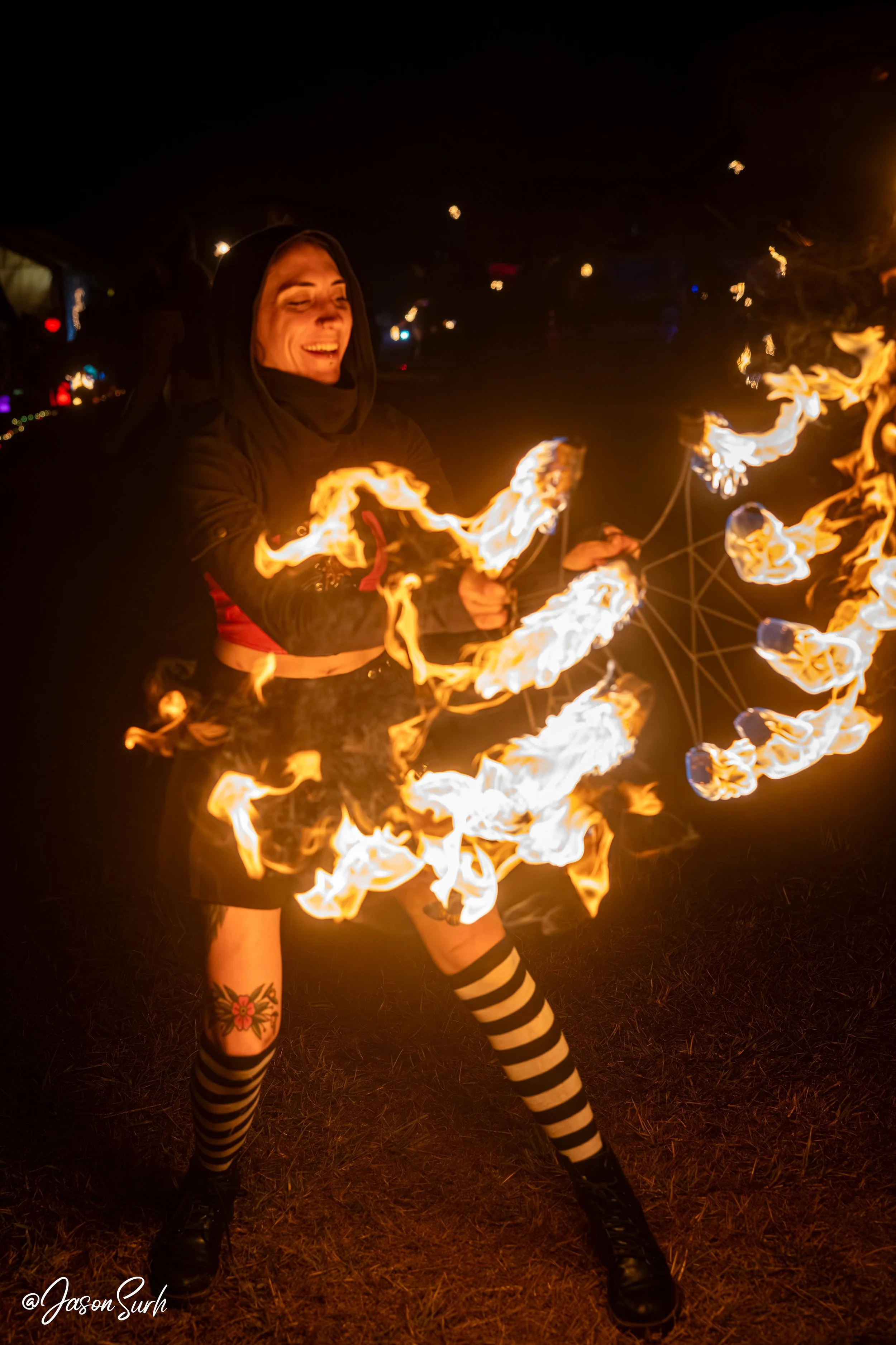 Person performing fire dancing at night, holding fire poi with flames around them, smiling, dressed in dark clothing with striped leggings and a tattoo on their thigh.