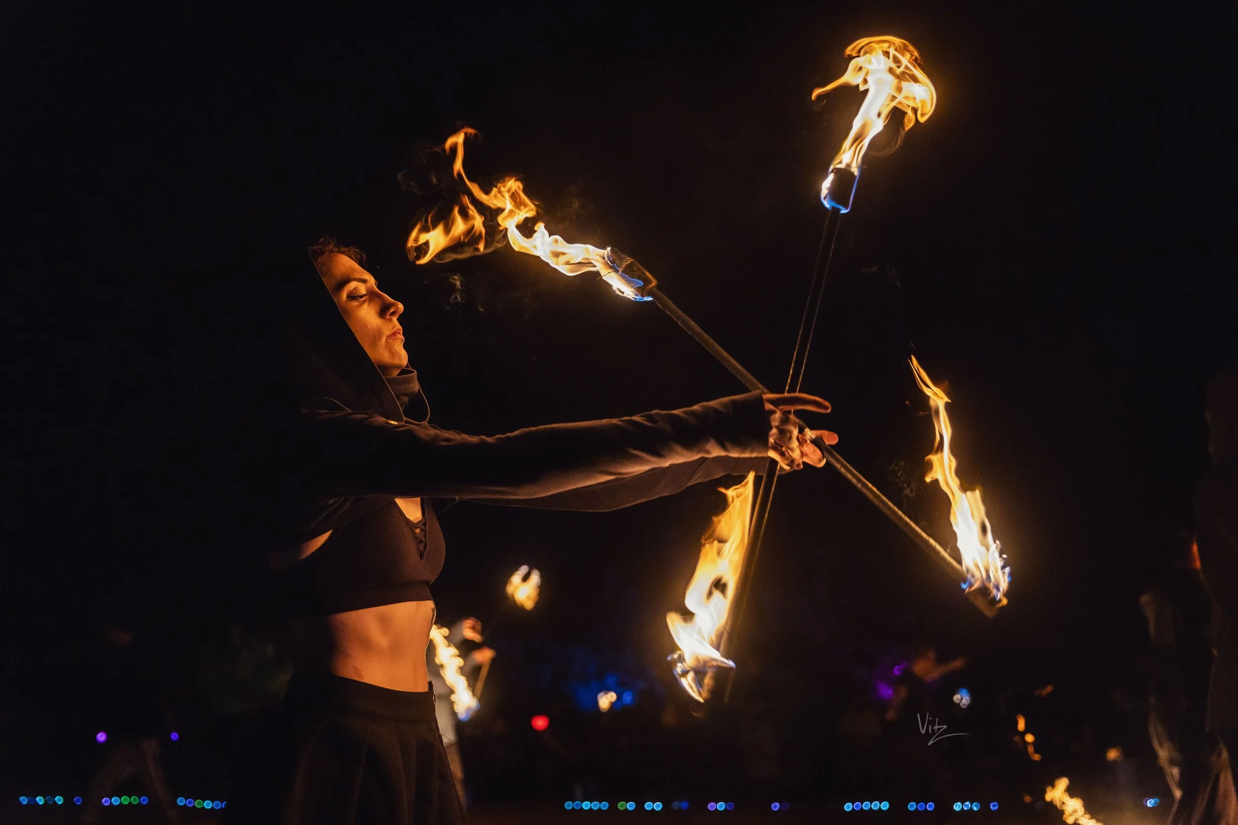 A woman performing a fire juggling act at night, with flames in each hand and on sticks, wearing a black outfit and hood.