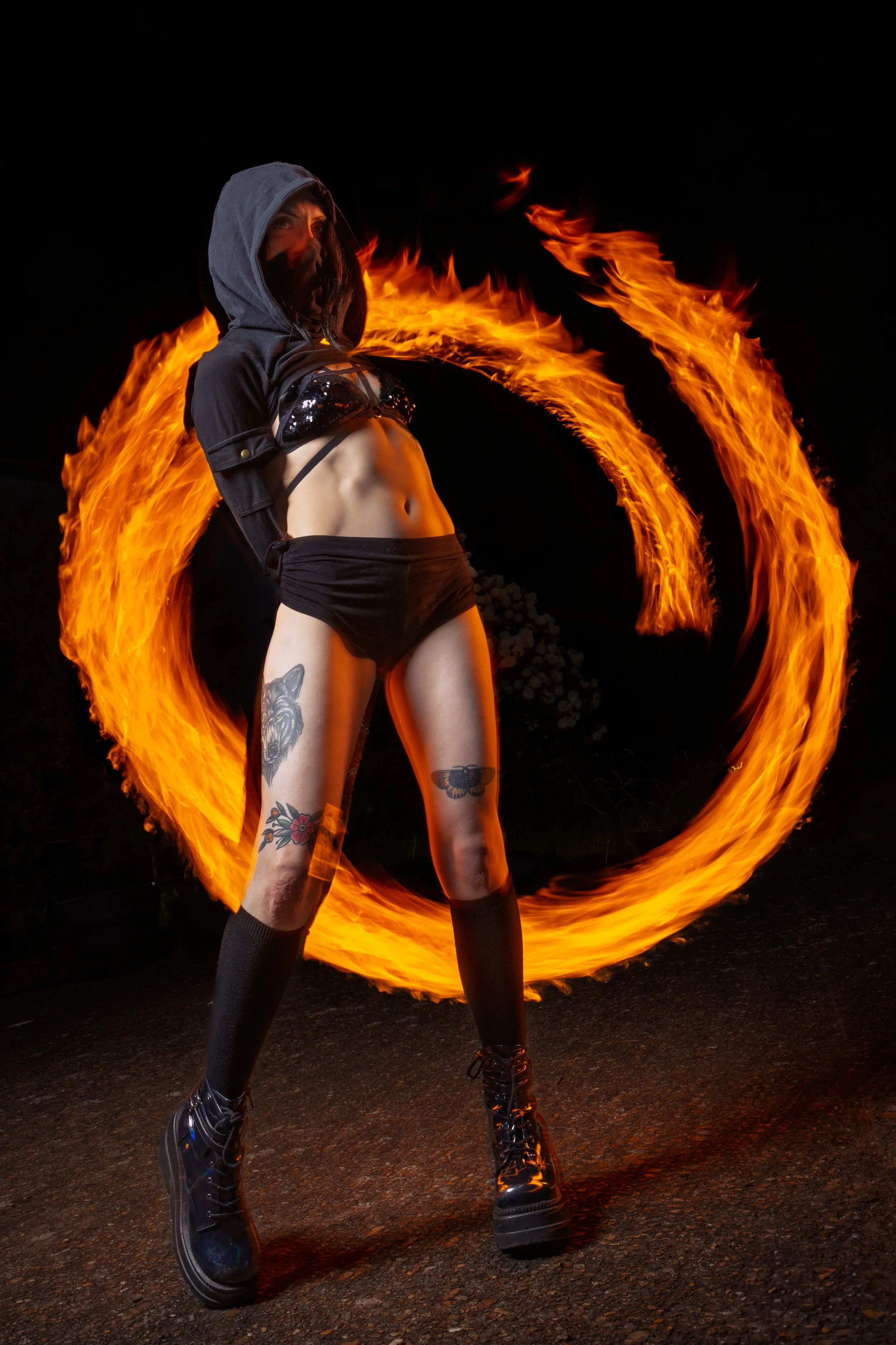 A woman performing fire spinning outdoors at night, creating a circle of flames with fire poi as she wears a hooded jacket, crop top, shorts, knee-high socks, and combat boots.