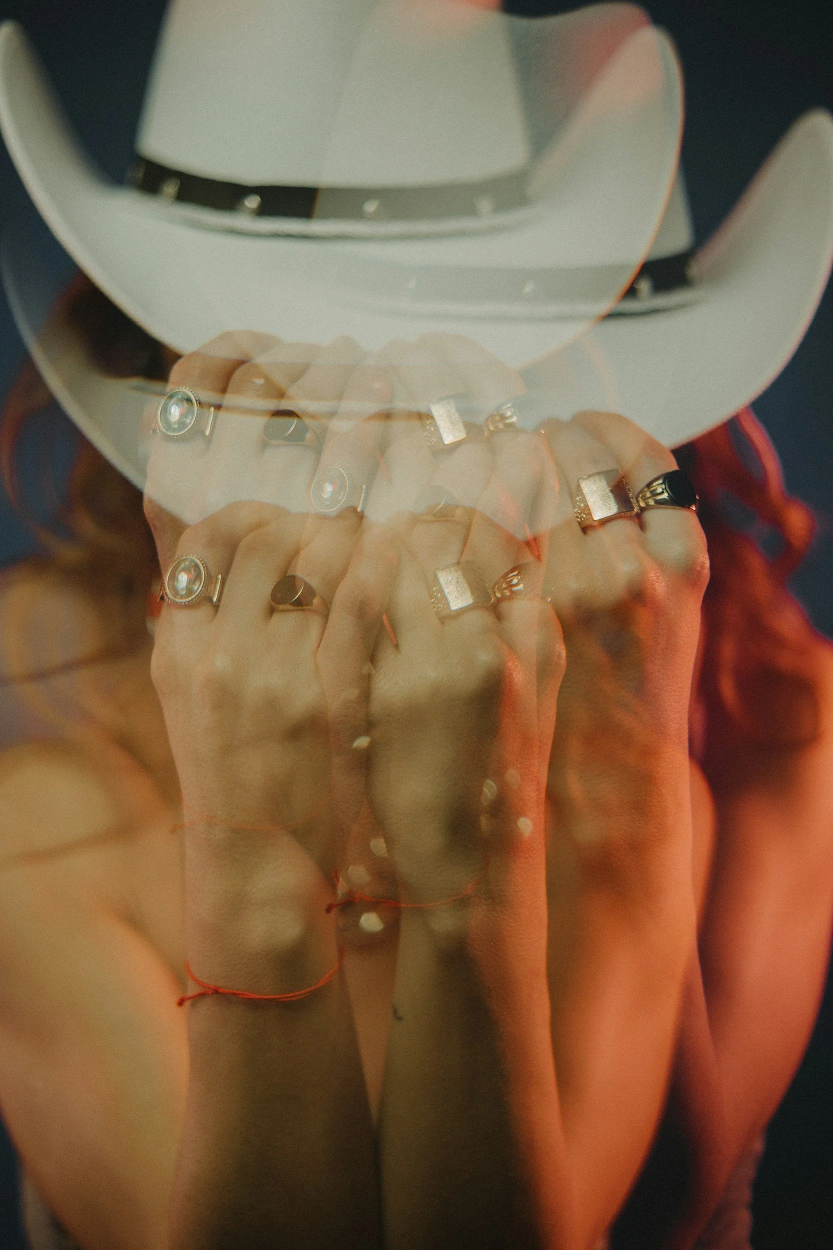 Close-up of a hand with multiple rings, outlined by warm lighting, overlaid with a faint reflection of a white wide-brimmed hat.