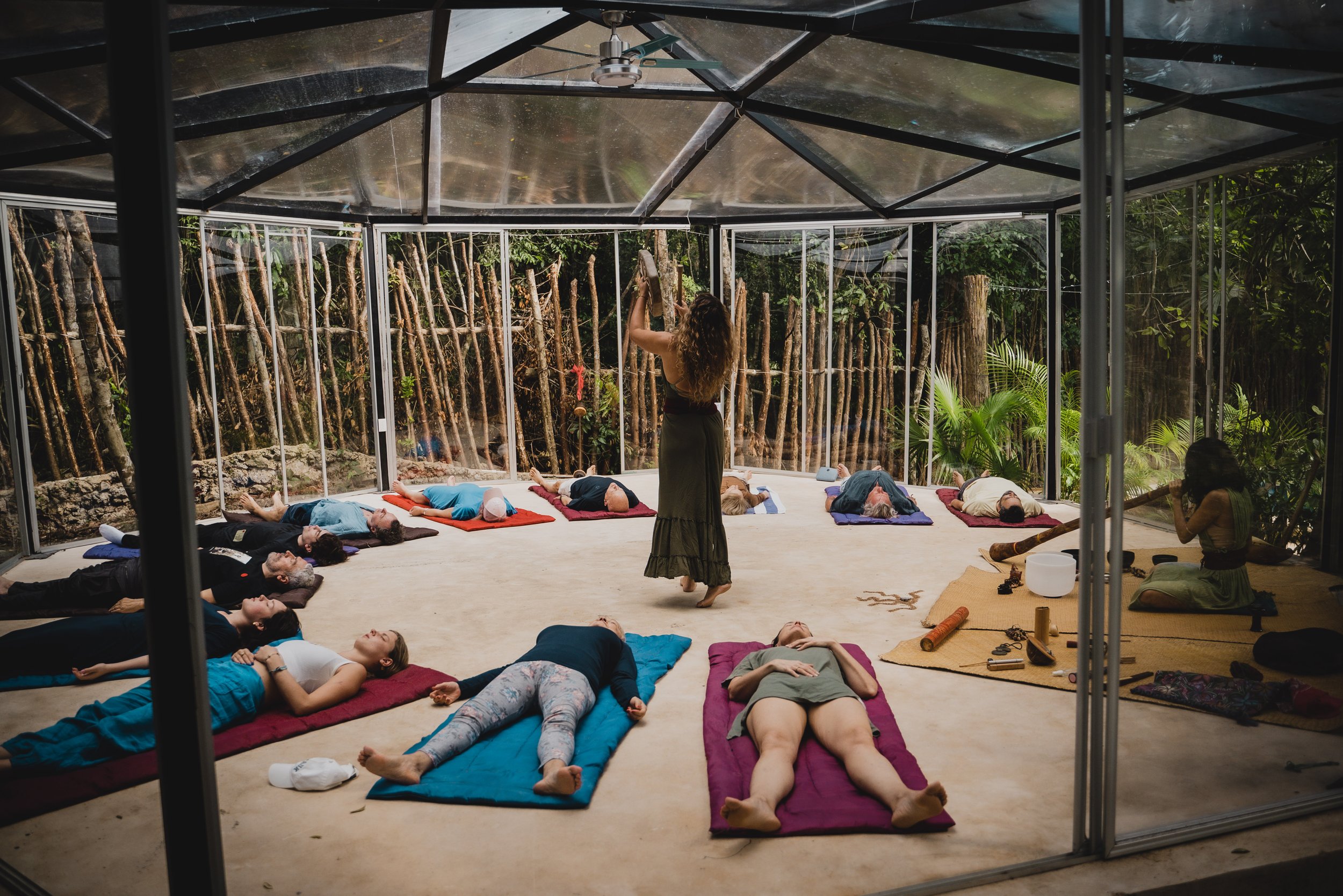 People lying on yoga mats in a glass-enclosed room while a woman leads a relaxation or meditation session; natural setting visible outside.