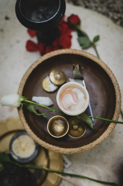 A table setting with a woven basket containing a white rose, open jars, and a seashell. A nearby gold tray holds additional items, and scattered red rose petals are visible.