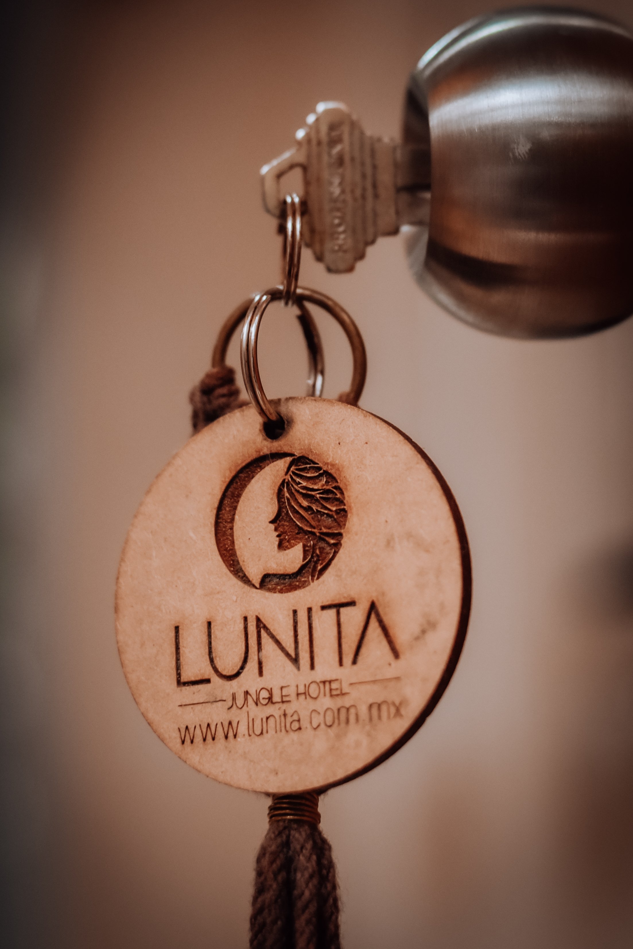 A door key with a wooden keychain engraved with "Lunita Jungle Hotel" logo, "www.lunita.com.mx," inserted into a metallic doorknob.