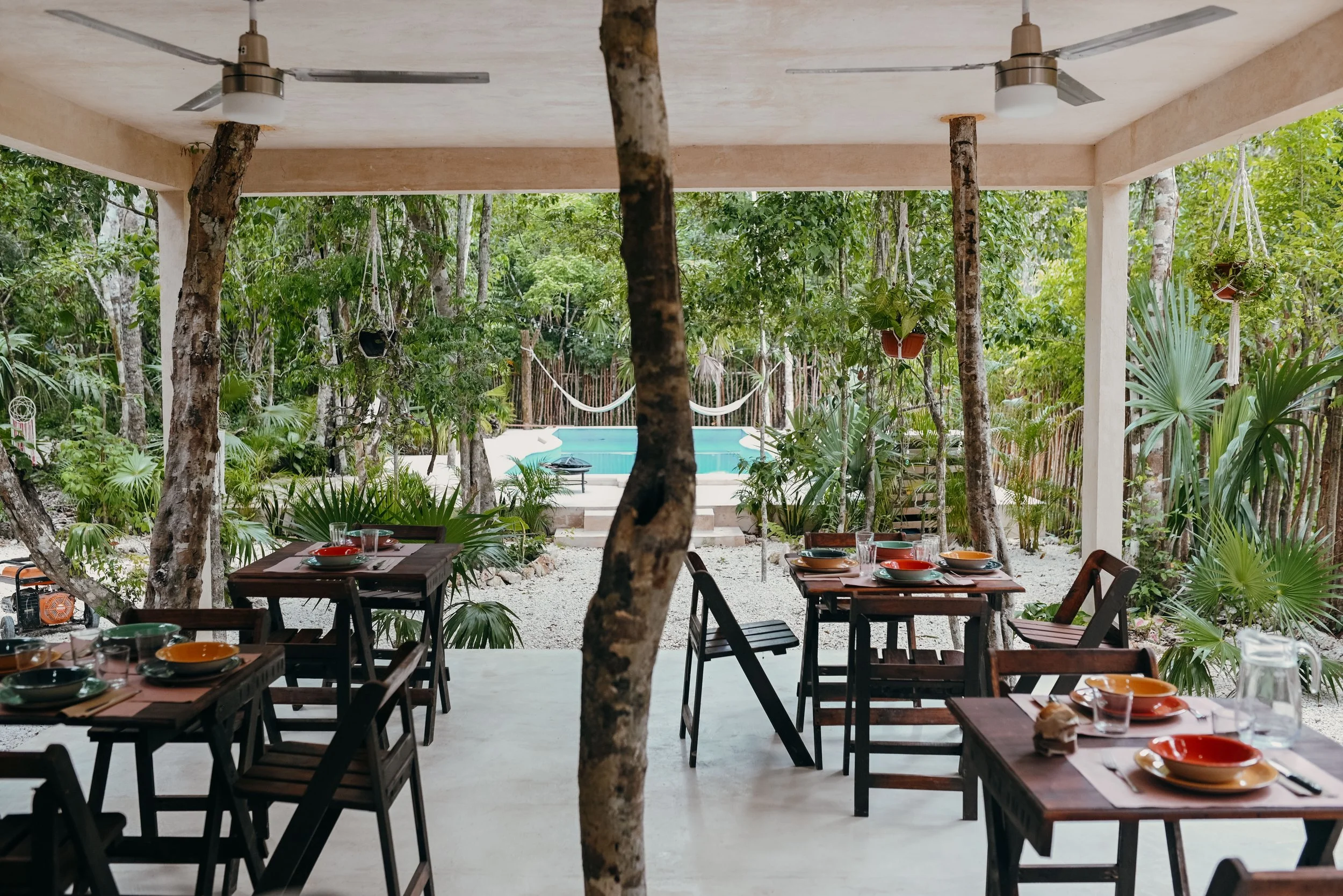 Outdoor dining area with wooden tables and chairs set for meals, surrounded by trees and plants, with a swimming pool and hammock in the background.