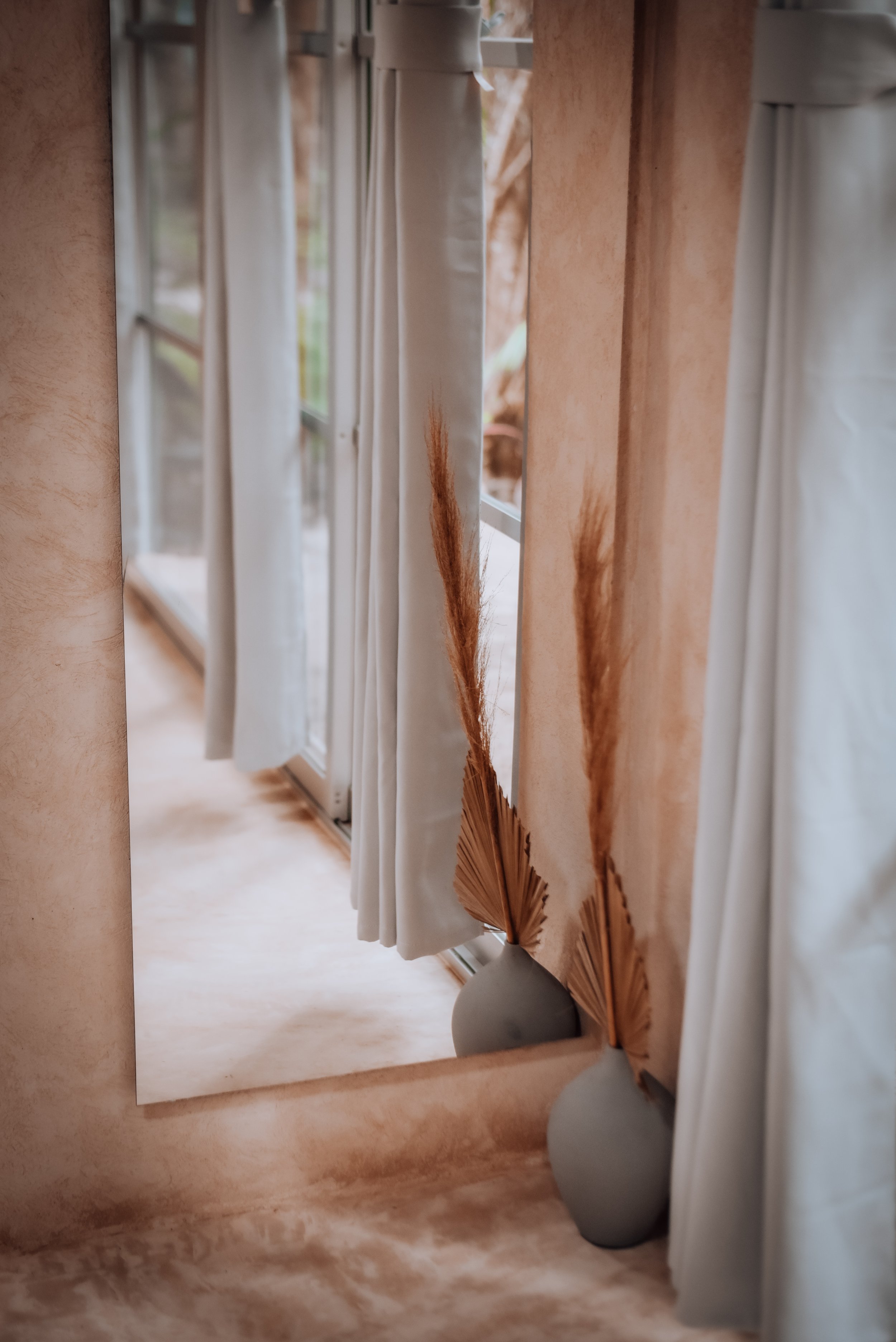 A room interior featuring a large window with sheer cream curtains. Below the window, there are two vases with tall, dried pampas grass and dried fan palm leaves, creating a minimalist and natural decor style. The room is softly lit, emphasizing the