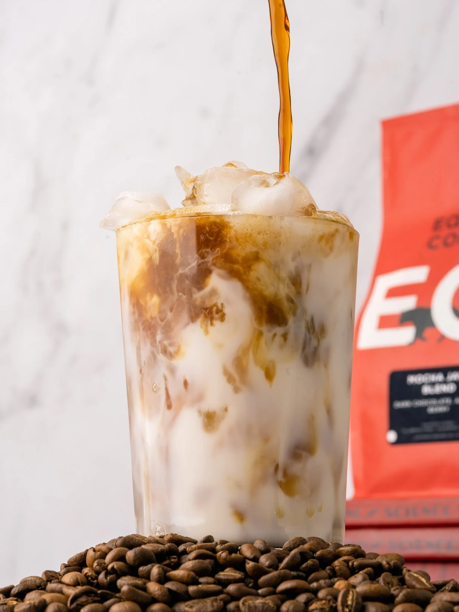 Iced coffee being poured into a glass. Equator coffee bag in the background and a mound of coffee beans is in the foreground