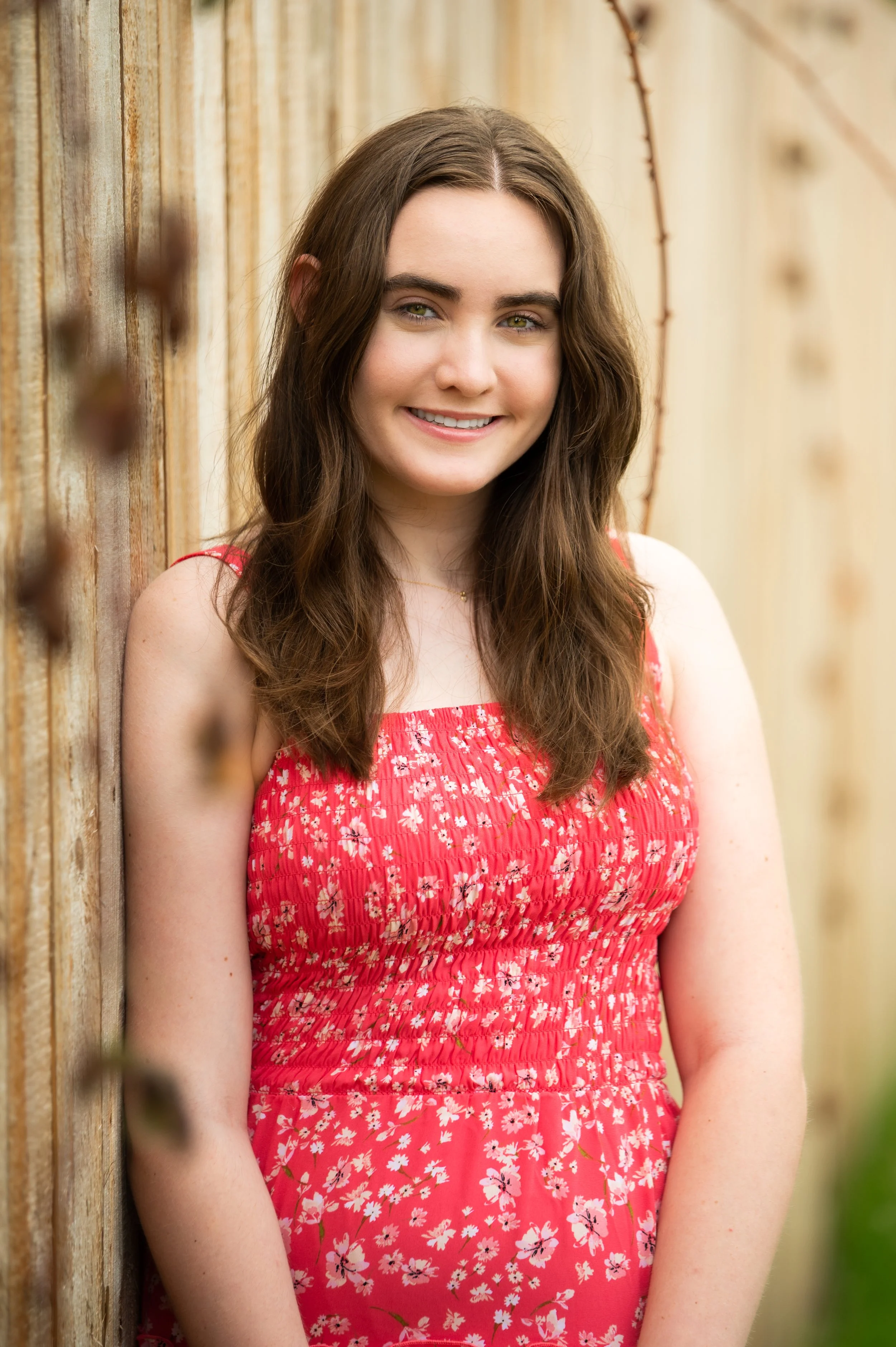 A young woman with long brown hair and green eyes, wearing a red sleeveless dress with white floral pattern, smiling and standing beside a wooden fence with some barbed wire.