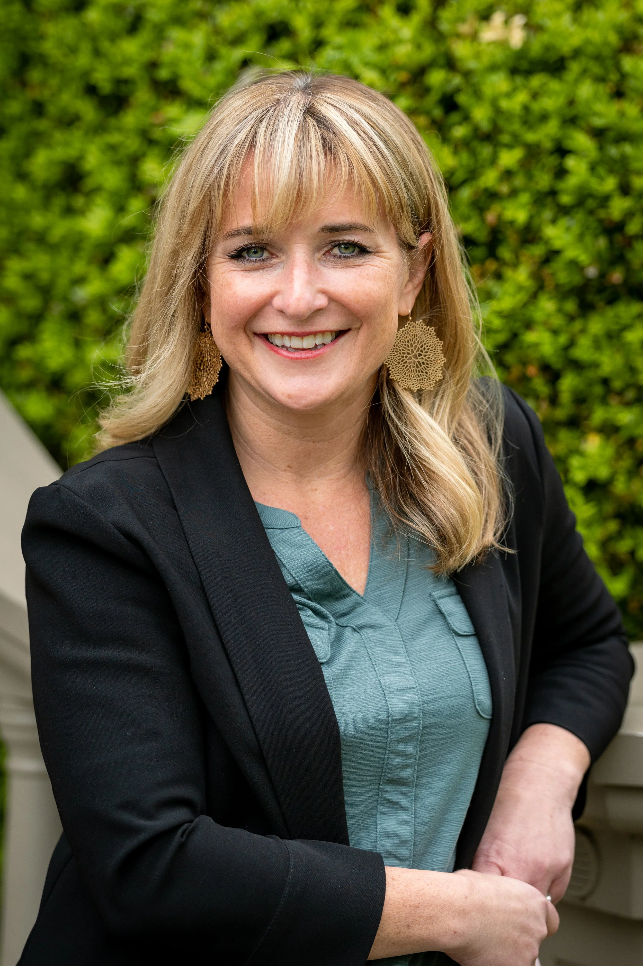 A woman with blonde hair smiling, wearing earrings and a black blazer over a teal blouse, standing outdoors in front of green shrubbery.