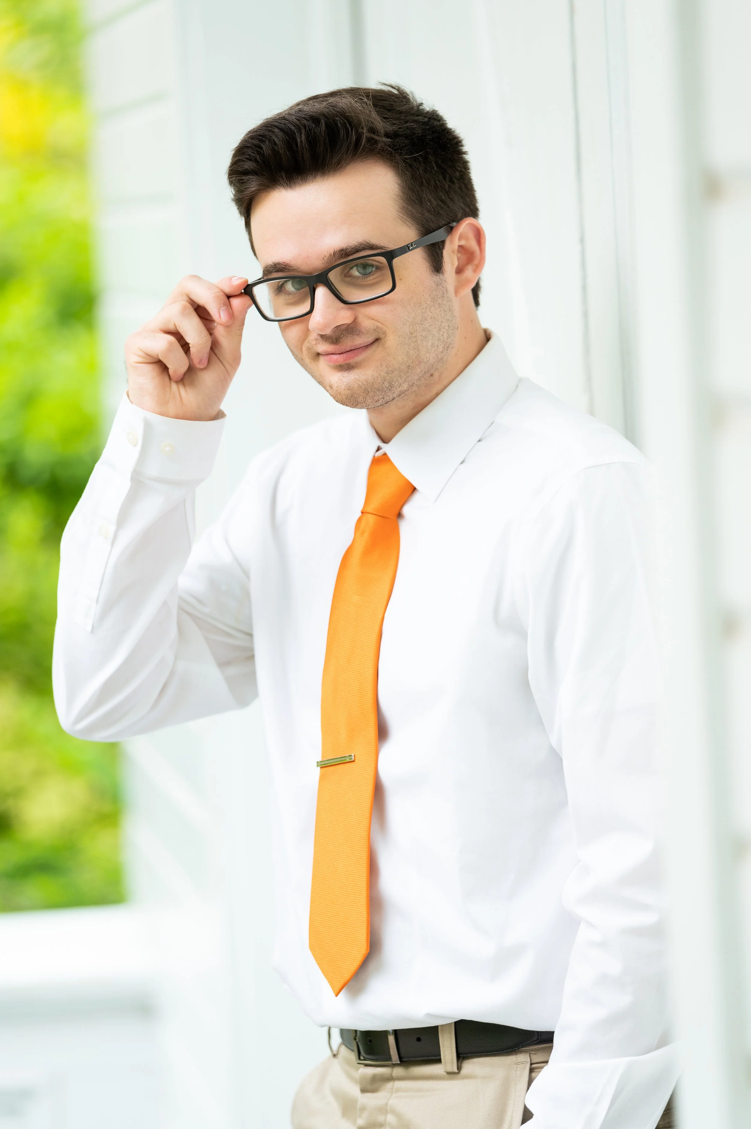 A young man in a white dress shirt, orange tie, and glasses adjusts his glasses while standing outside near a white building with greenery in the background.