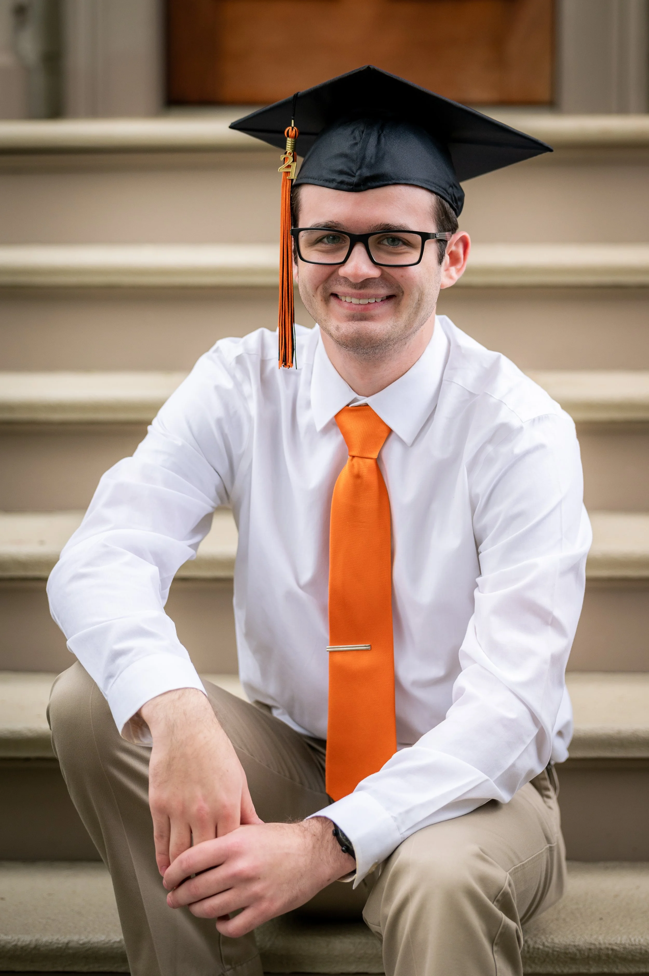 A young man wearing black graduation cap with orange tassel, glasses, white shirt, orange tie, and beige pants, sitting on stairs and smiling at the camera.