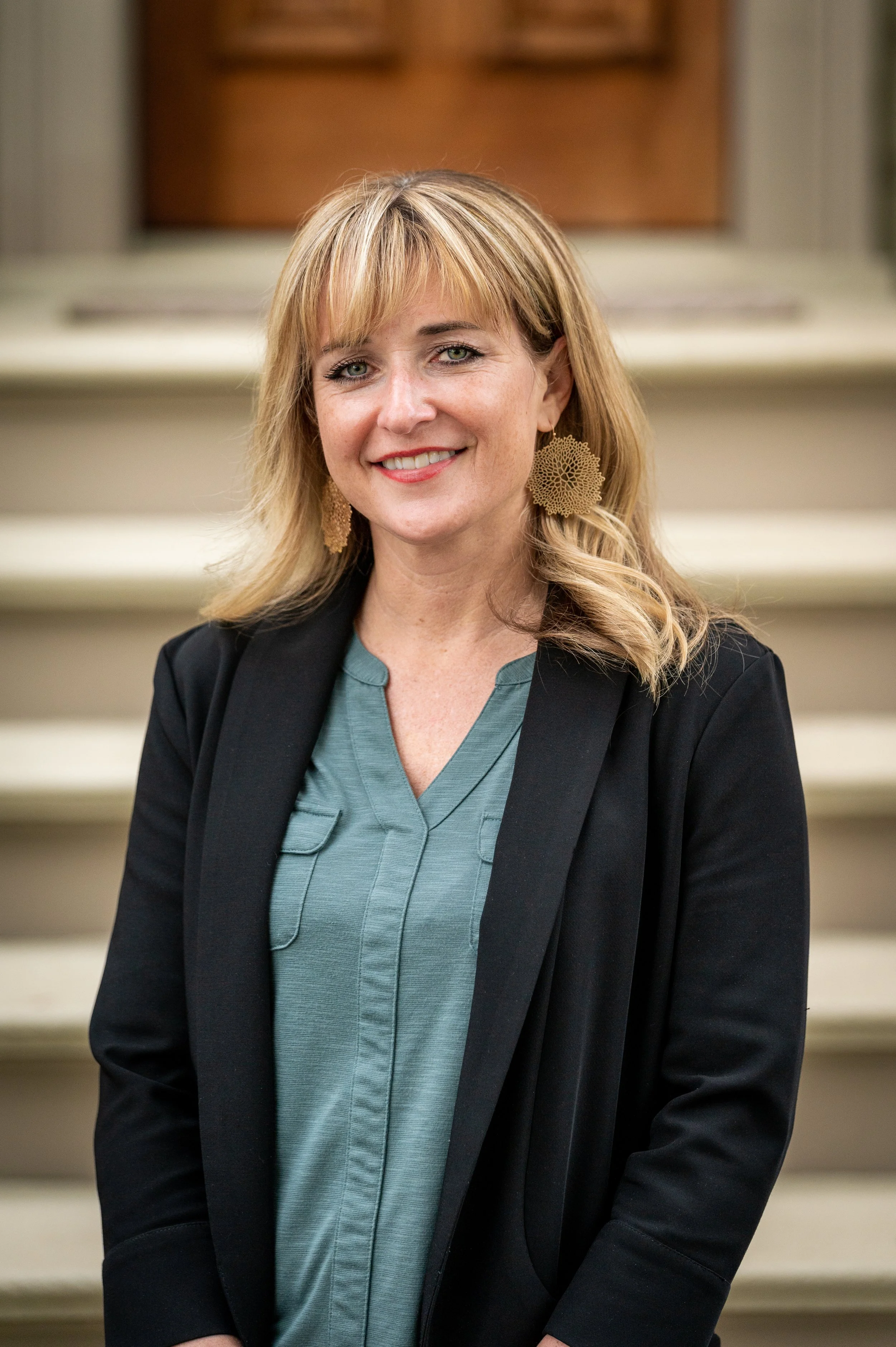 A woman with blonde hair, wearing a black blazer and teal blouse, smiling, standing in front of stairs.